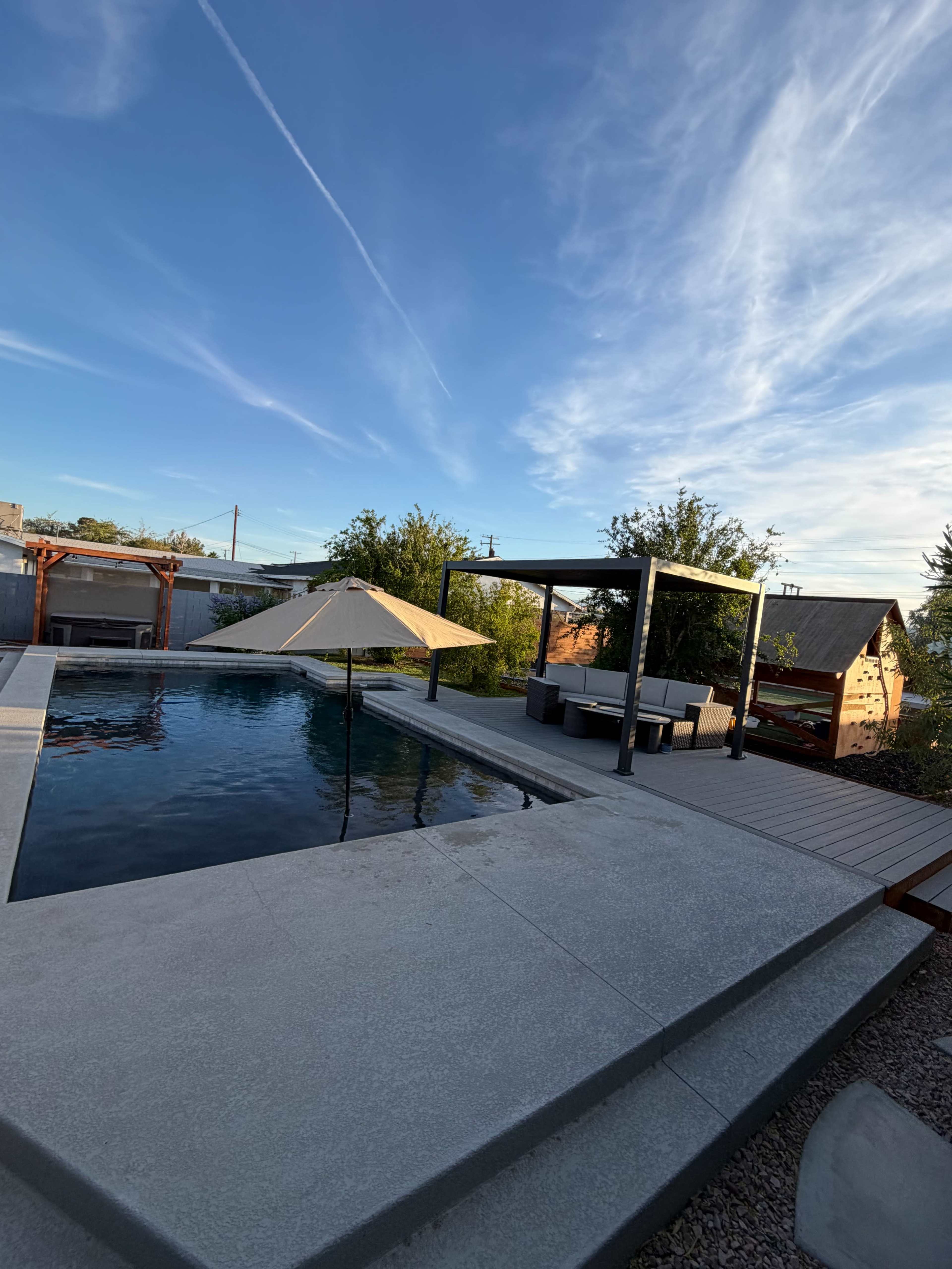 The image shows a modern backyard featuring a swimming pool with a shaded seating area, surrounded by landscaping and a clear blue sky.