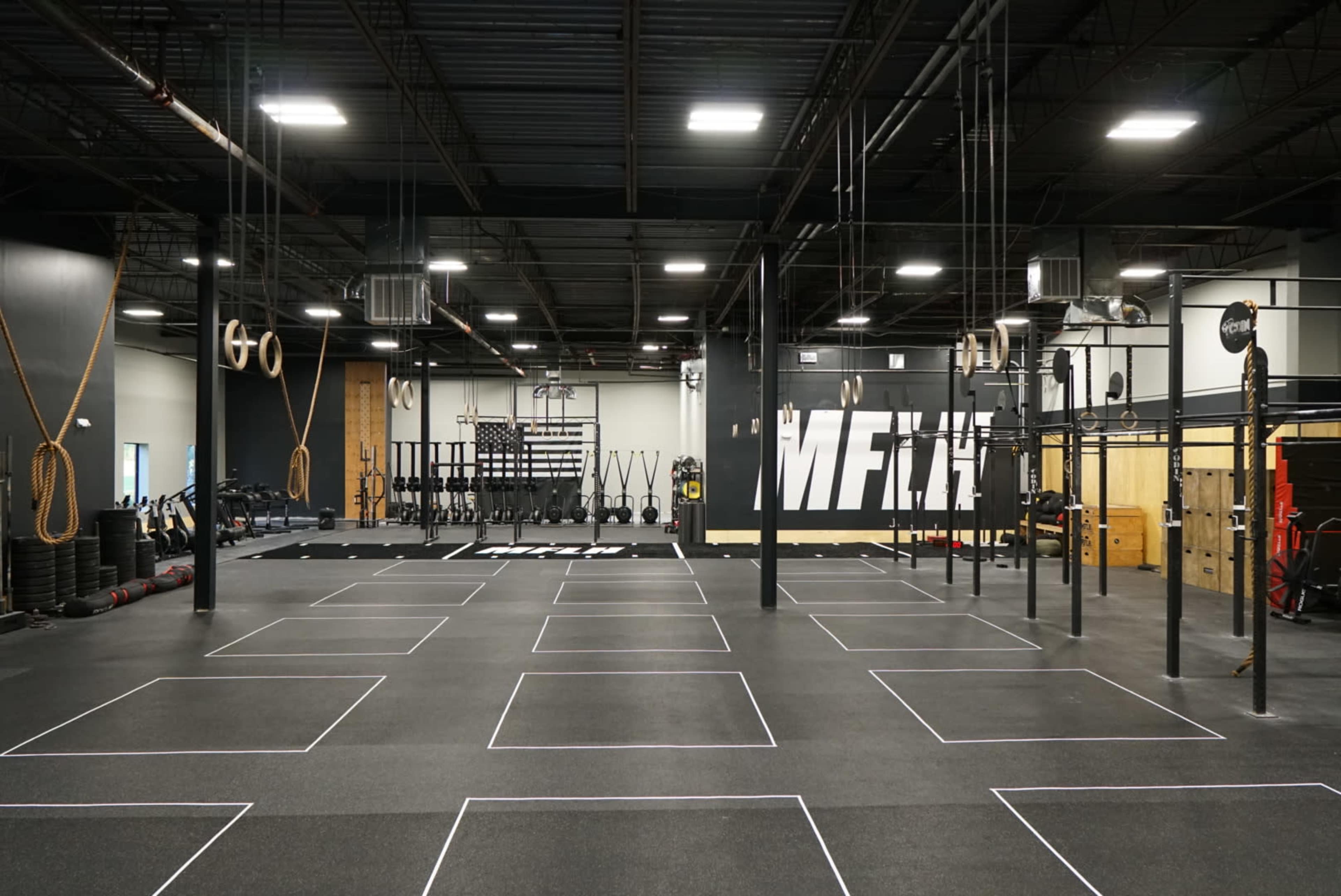 The image shows a spacious gym interior with various workout equipment, marked exercise zones, and a large wall mural that reads "MFT."