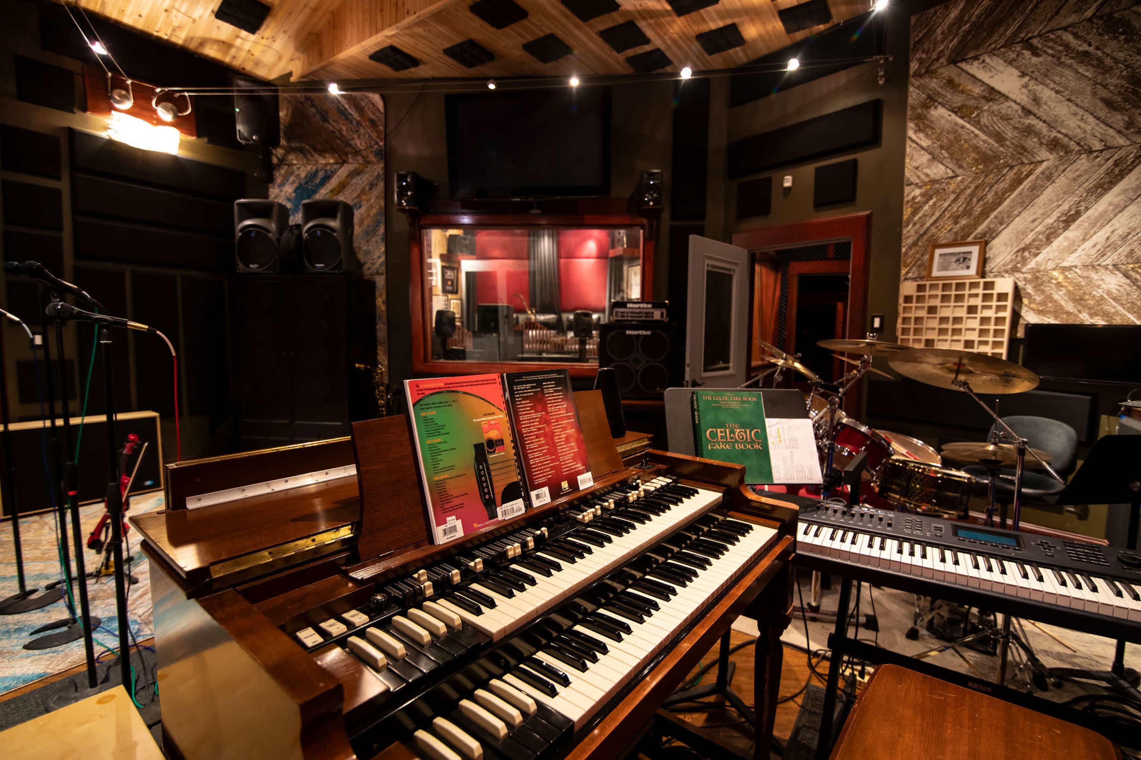 A recording studio filled with various musical instruments, including an organ, keyboard, and a drum set, along with sound equipment and a soundproof booth in the background.