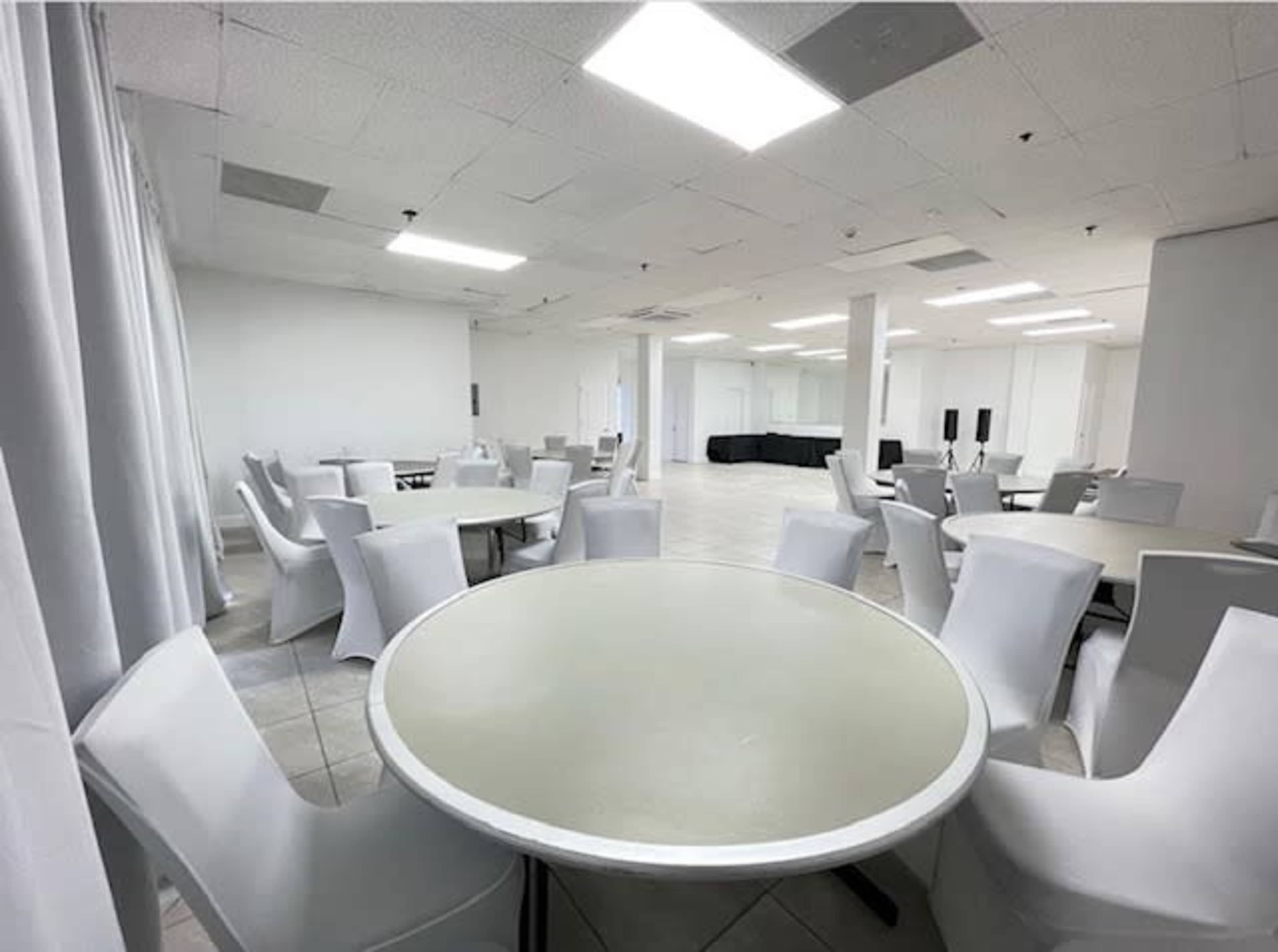 The image shows a spacious, empty room with several round tables surrounded by white chairs and bright overhead lights.