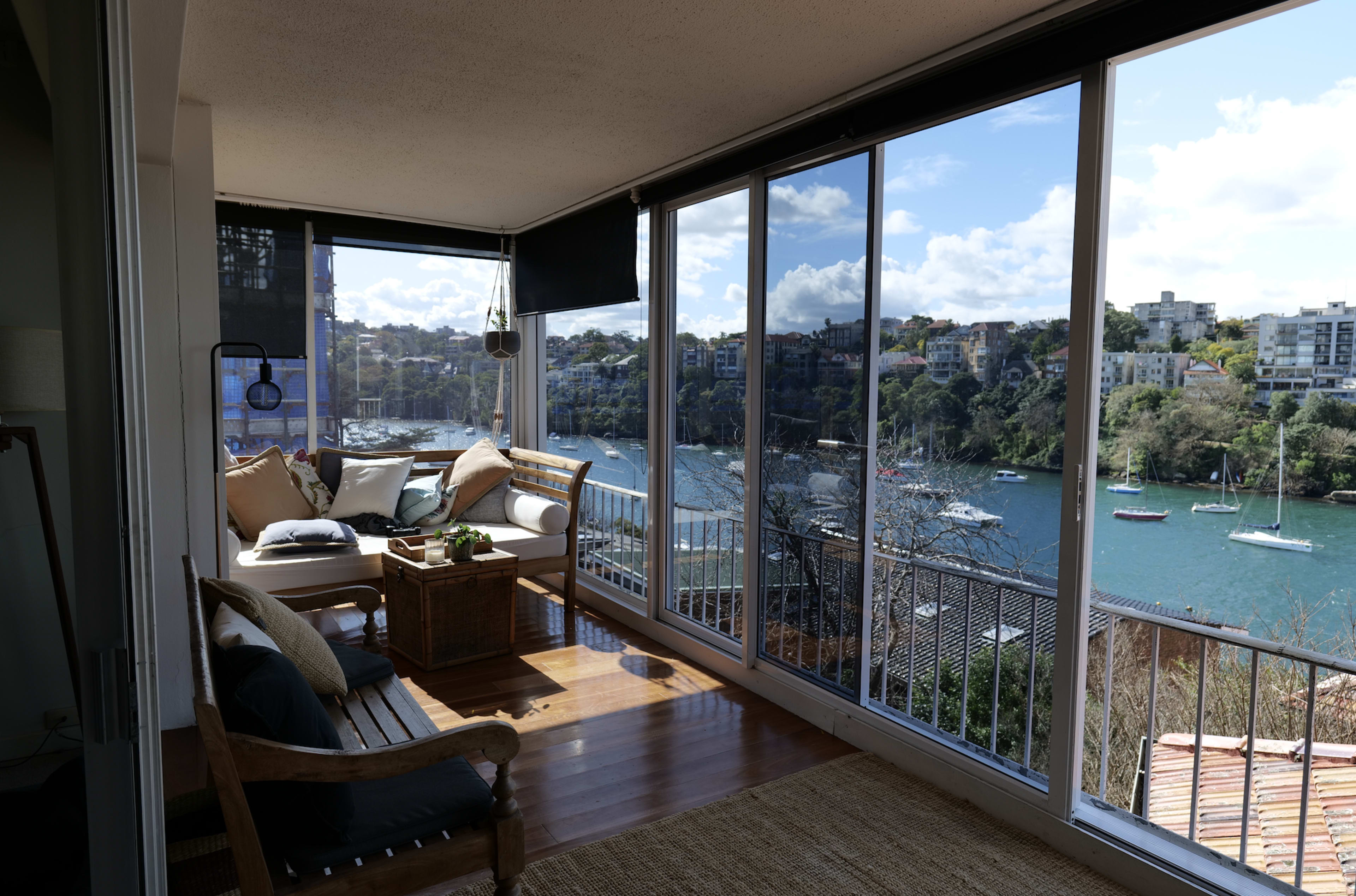 The image shows a sunlit balcony with a seating area overlooking a harbor dotted with boats and framed by trees and buildings.