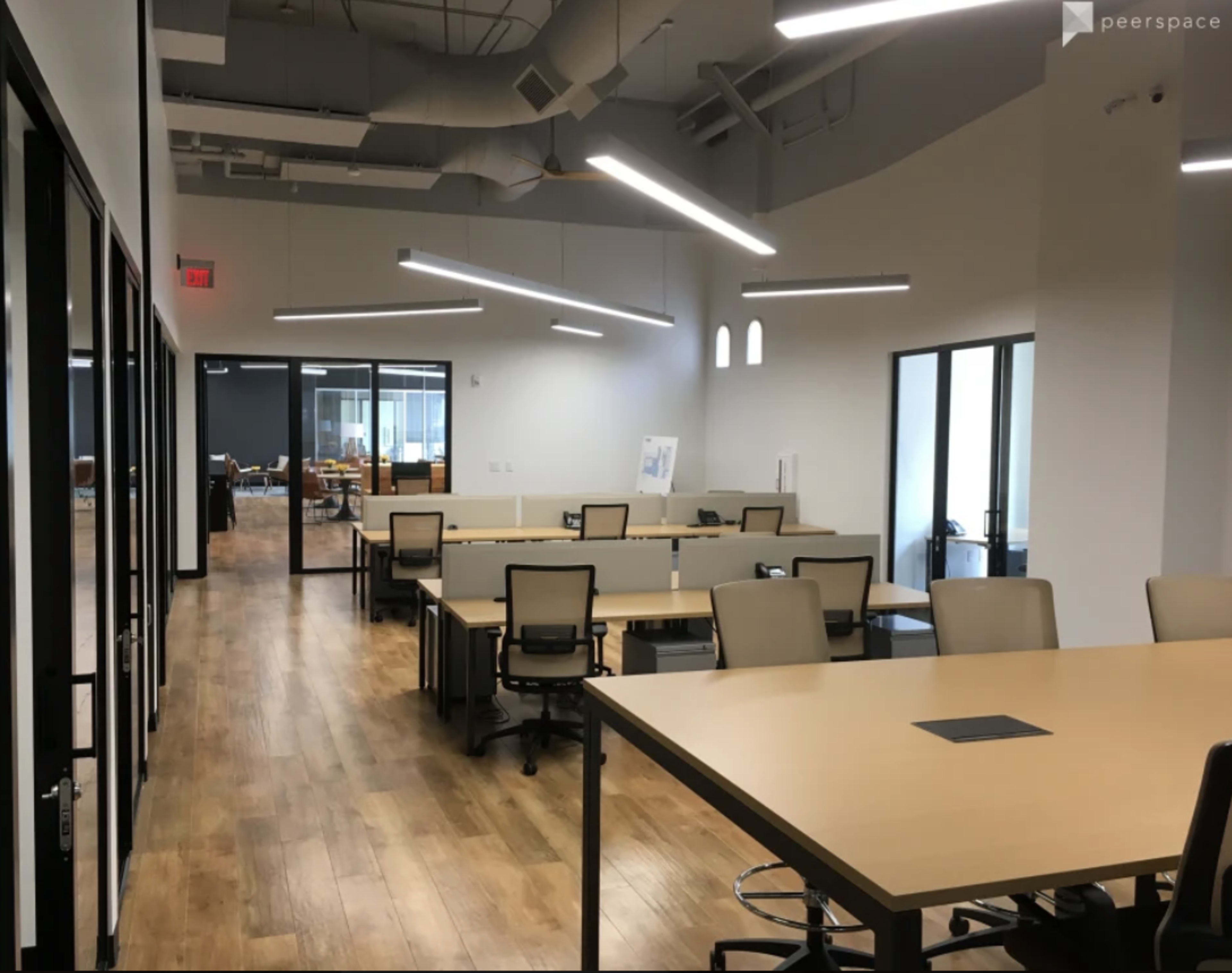 A modern office space with multiple desks arranged in a long row, large windows, and industrial-style lighting.