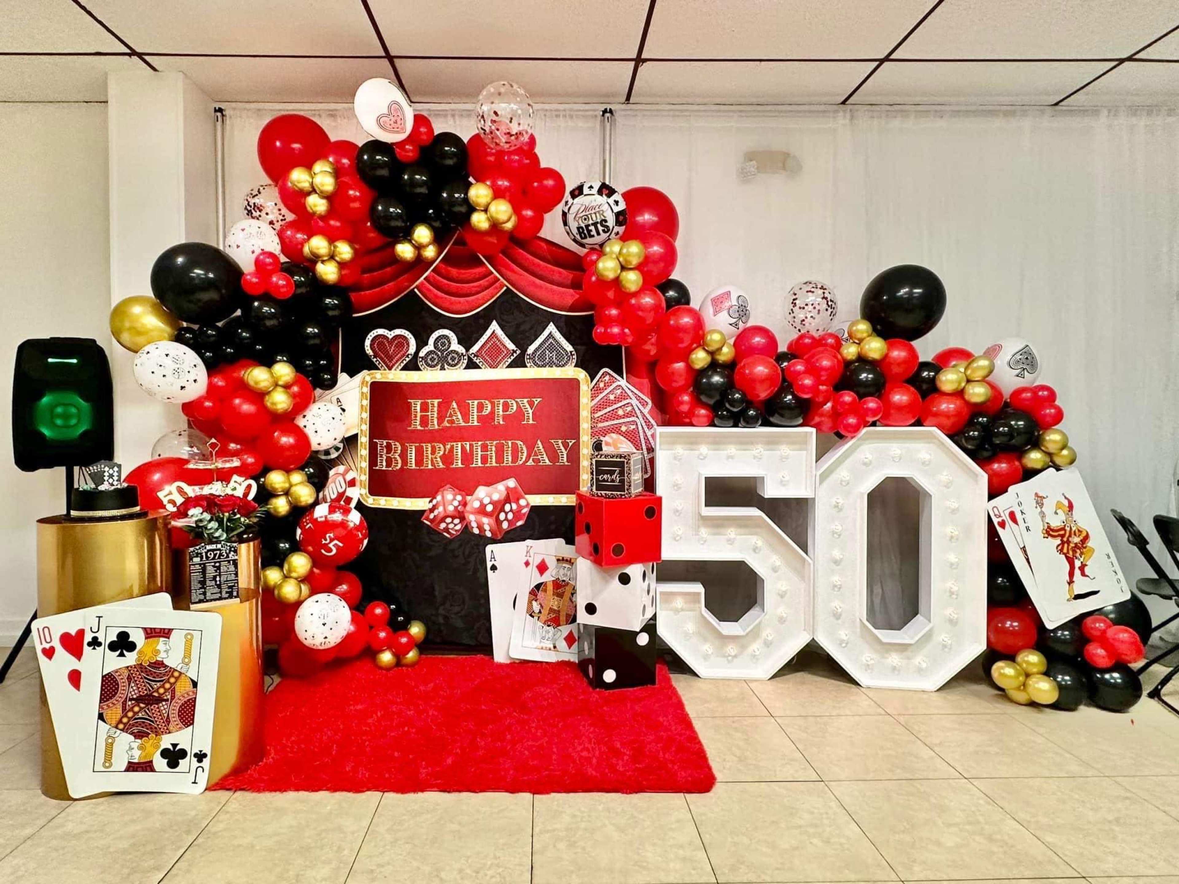 The image shows a festive birthday party setup featuring red, black, and gold balloons, a large "50" sign, and decorative elements like playing cards and a dice.