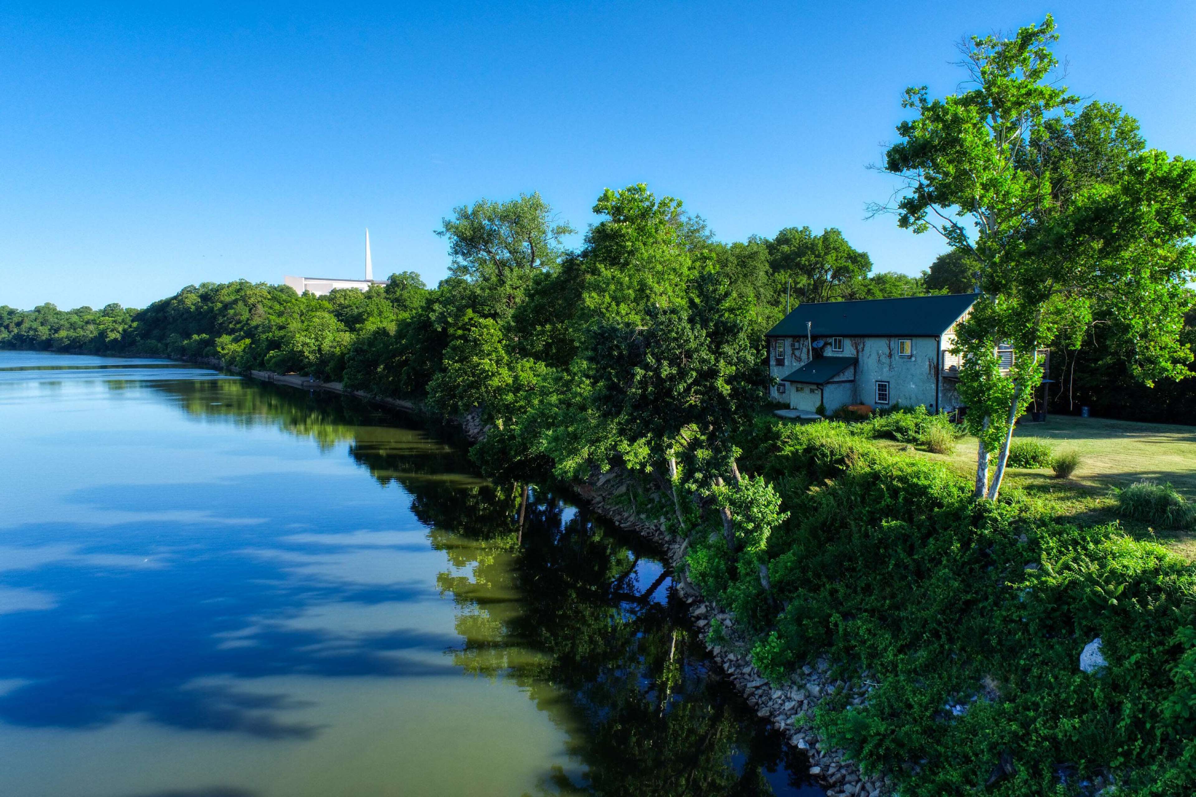 A house with a green roof is situated on the bank of a calm river, surrounded by lush trees and vegetation.