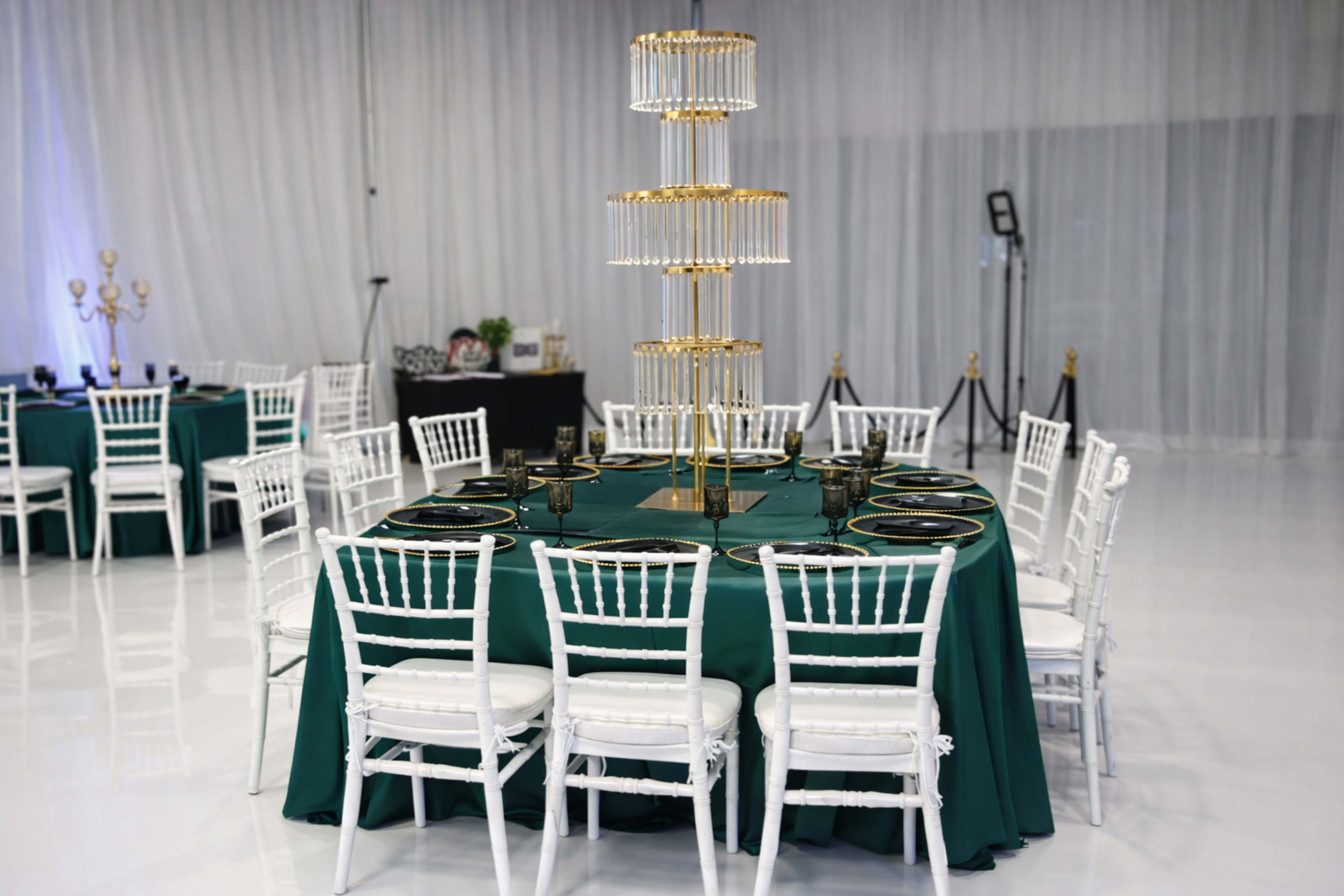 A round dining table with a green tablecloth is set with black plates and surrounded by white chiavari chairs, featuring a tall, gold candle holder centerpiece in a spacious event space.