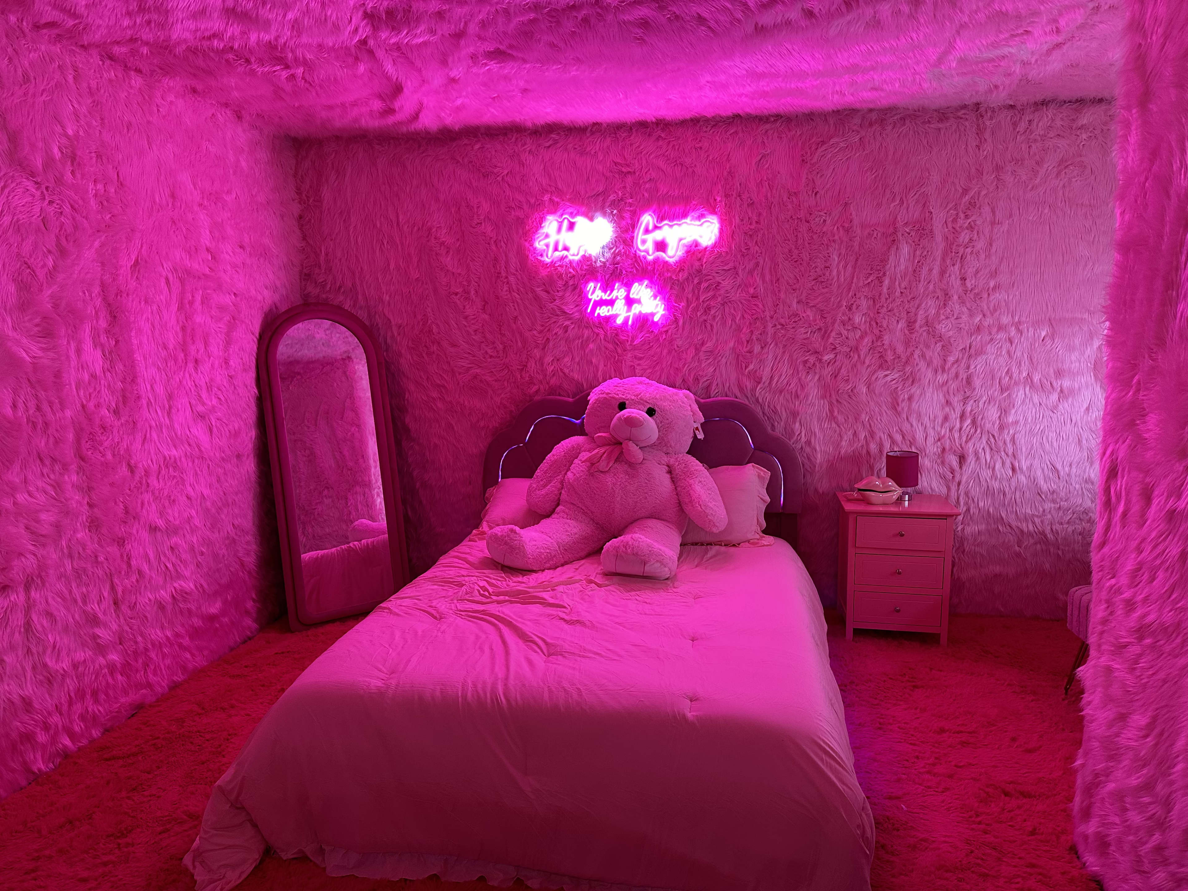 The room is entirely covered in pink fur, featuring a large pink teddy bear on the bed, neon signs on the wall, and a mirrored dresser.