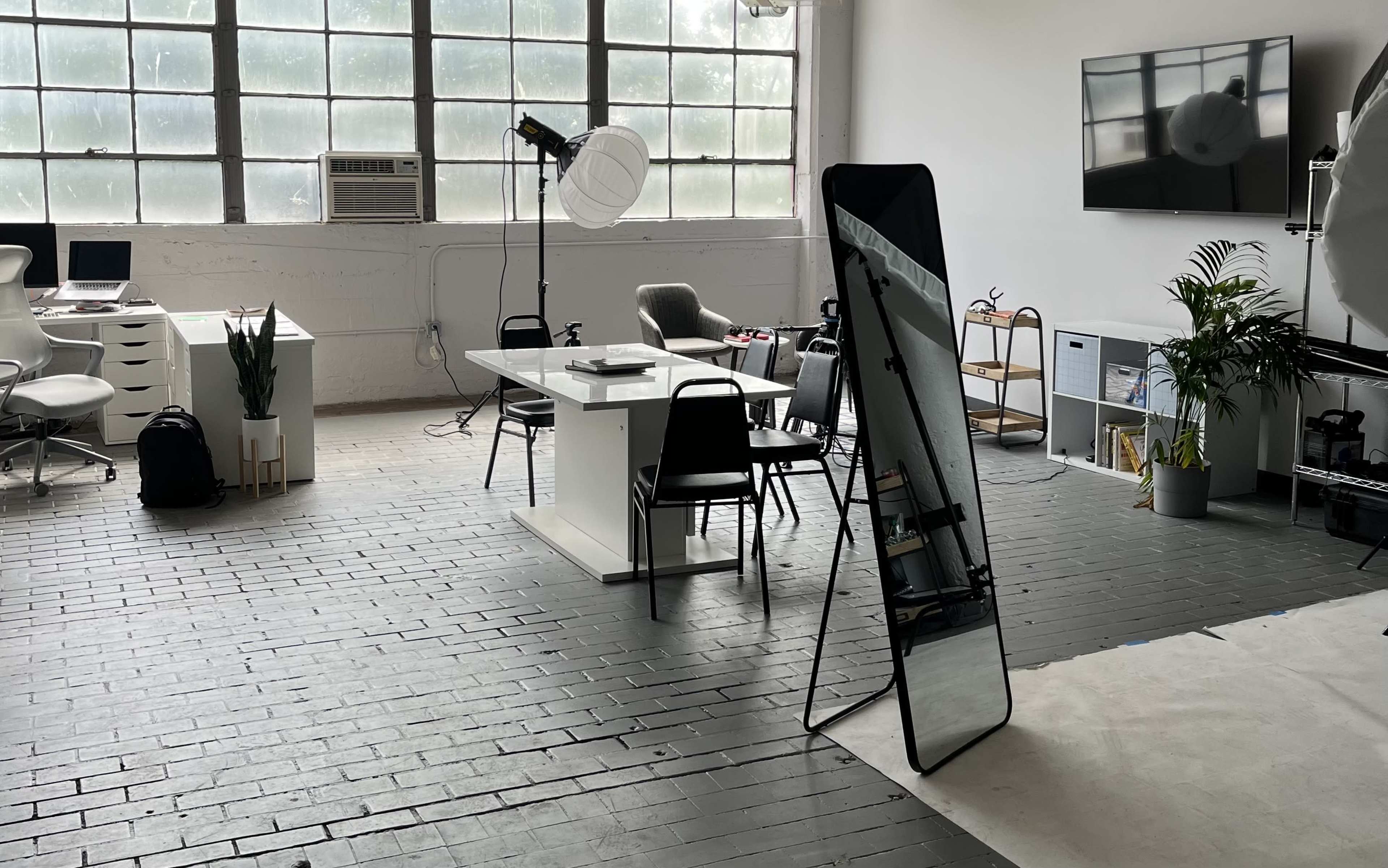 A spacious office environment featuring a large window, a white desk with chairs, an oversized mirror, and various plants.