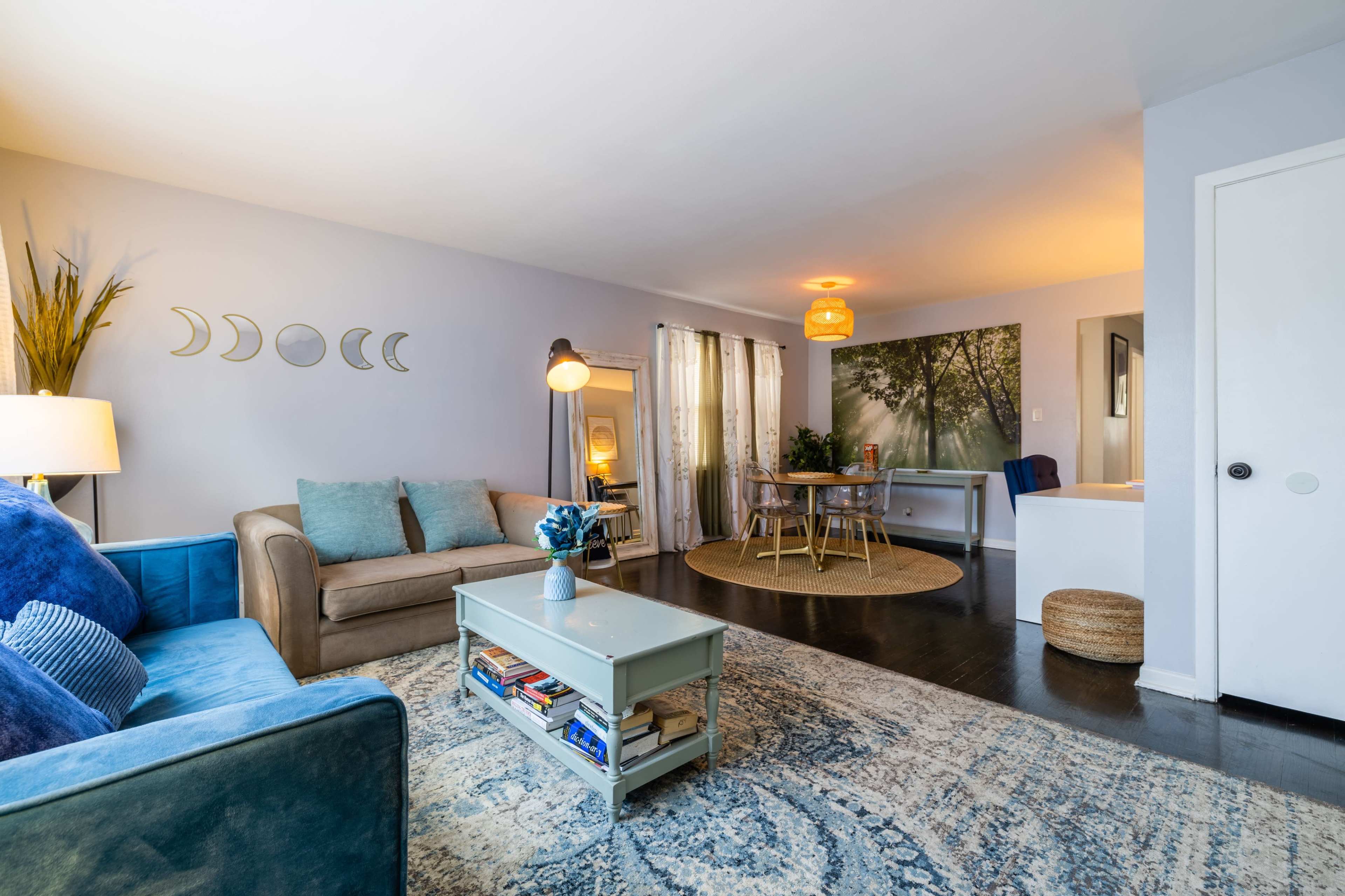 The image shows a living room with a blue sofa, a coffee table stacked with books, and a dining area featuring a round table and greenery, all set against light gray walls.