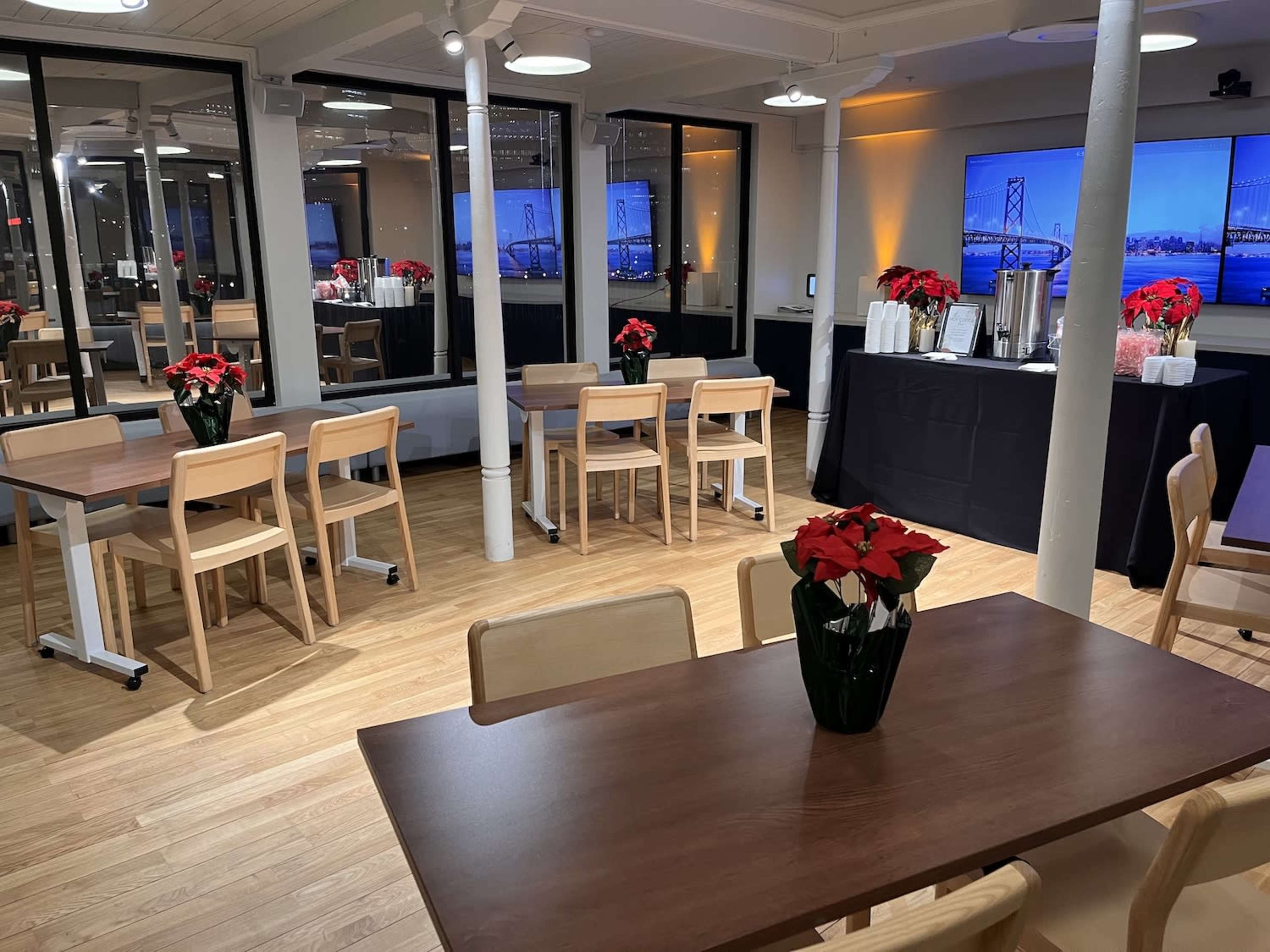 The image shows a modern event space with wooden floors, tables set for dining, and a refreshment station in the background featuring a large screen displaying a cityscape.