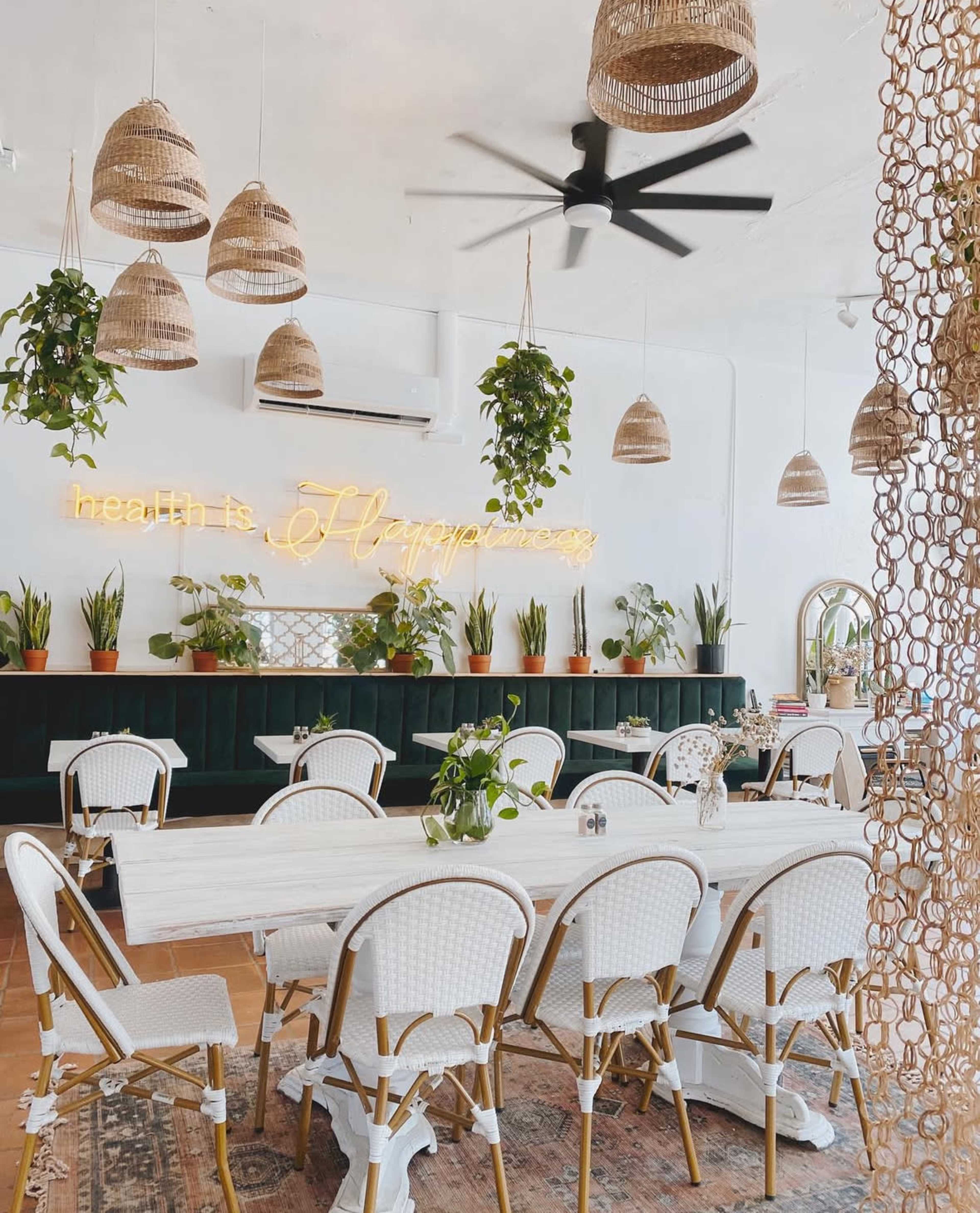 The image shows a bright dining area with a long white table surrounded by white wicker chairs, decorated with hanging plants and rattan light fixtures.
