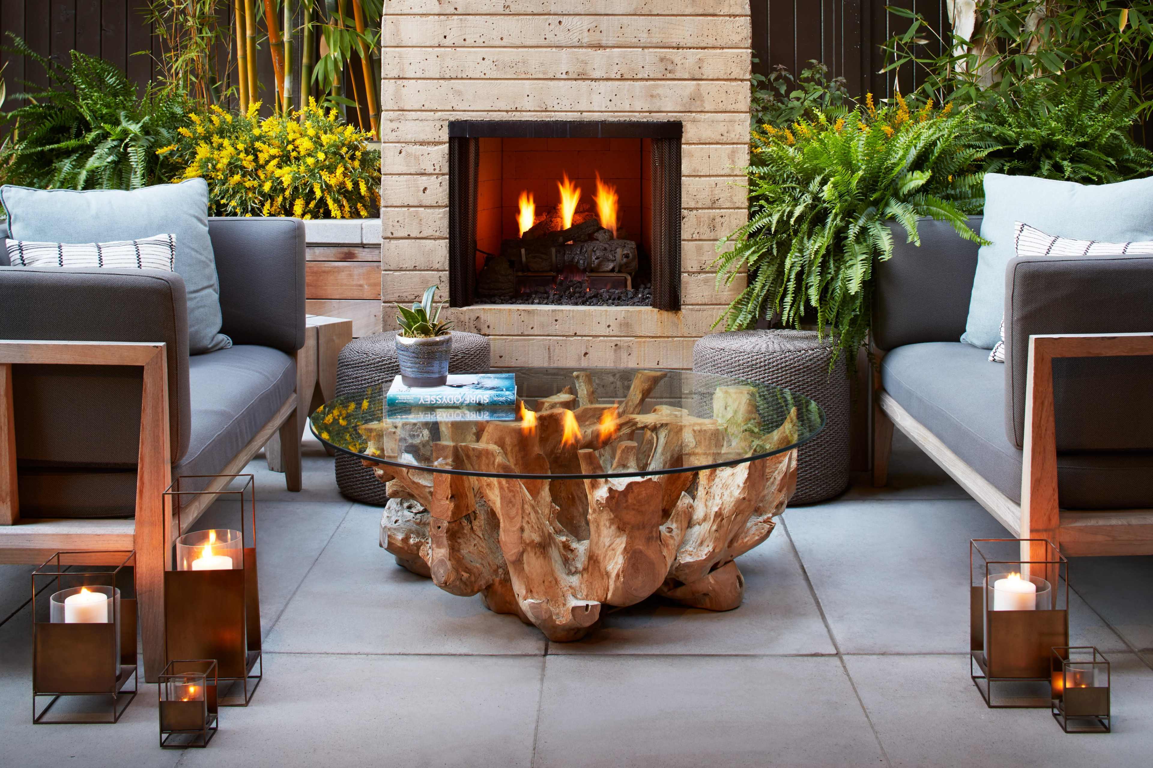 A cozy outdoor seating area with two armchairs, a wooden coffee table, and a fireplace surrounded by greenery and decorative lanterns.