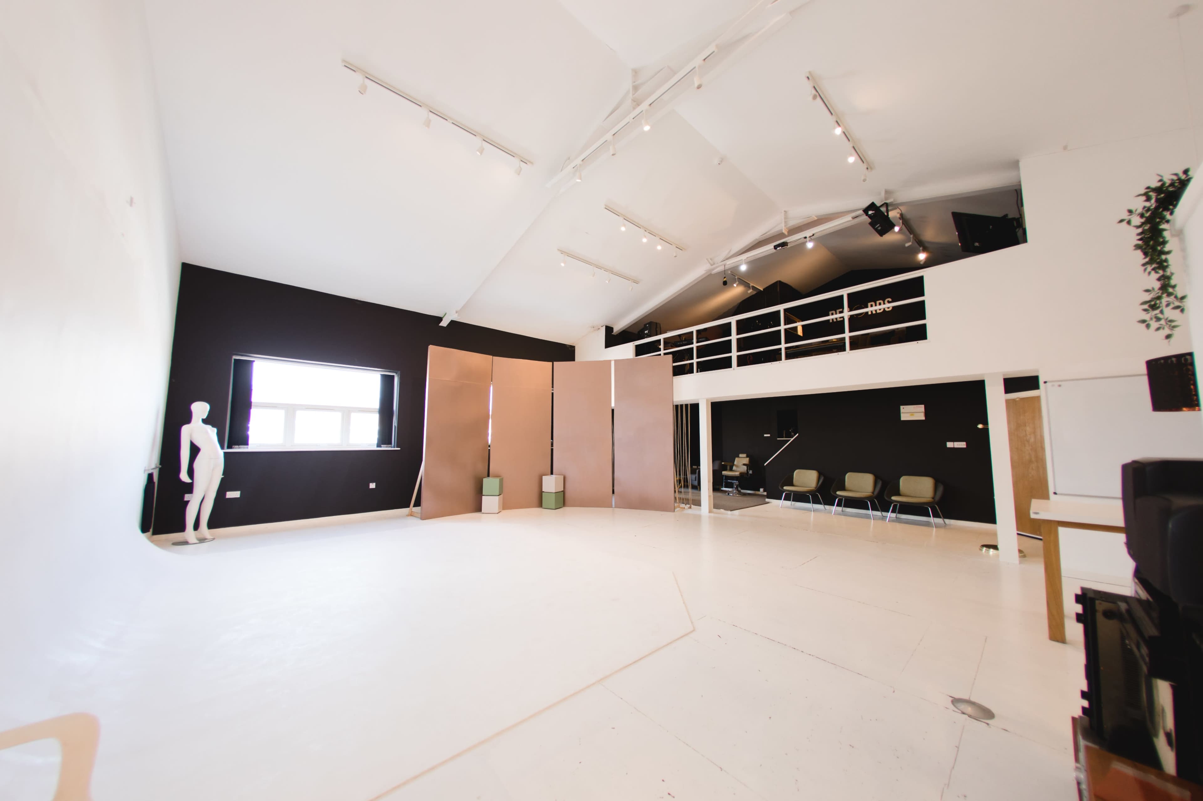 A spacious studio with white floors, black walls, a large window, and a loft area featuring a few chairs and a mannequin in the foreground.