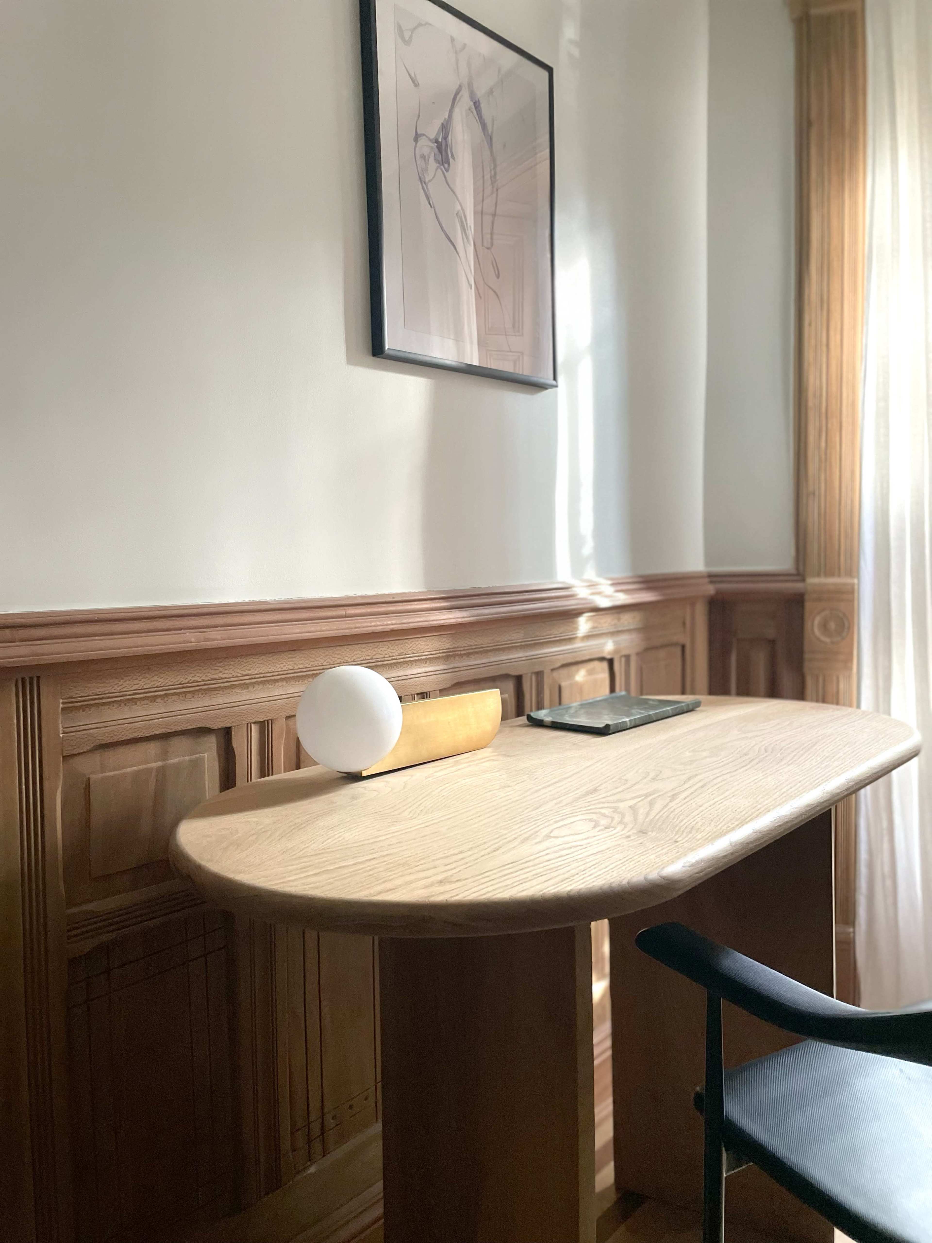 A minimalist wooden desk with a rounded edge, a round lamp, and a tablet rests next to a wall adorned with wood paneling and a framed artwork.