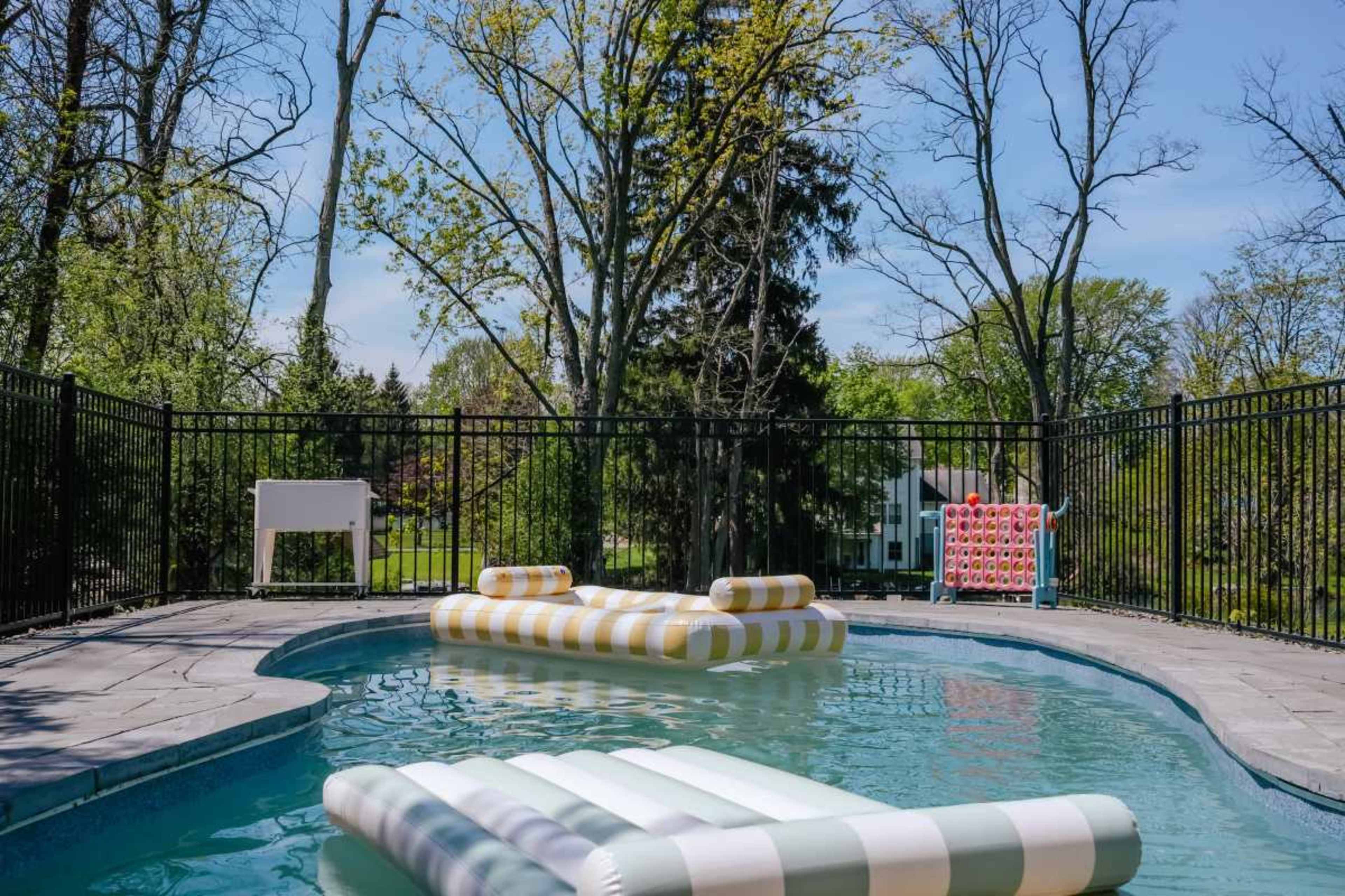 A swimming pool features two striped inflatable loungers floating on the water, surrounded by trees and a fence.