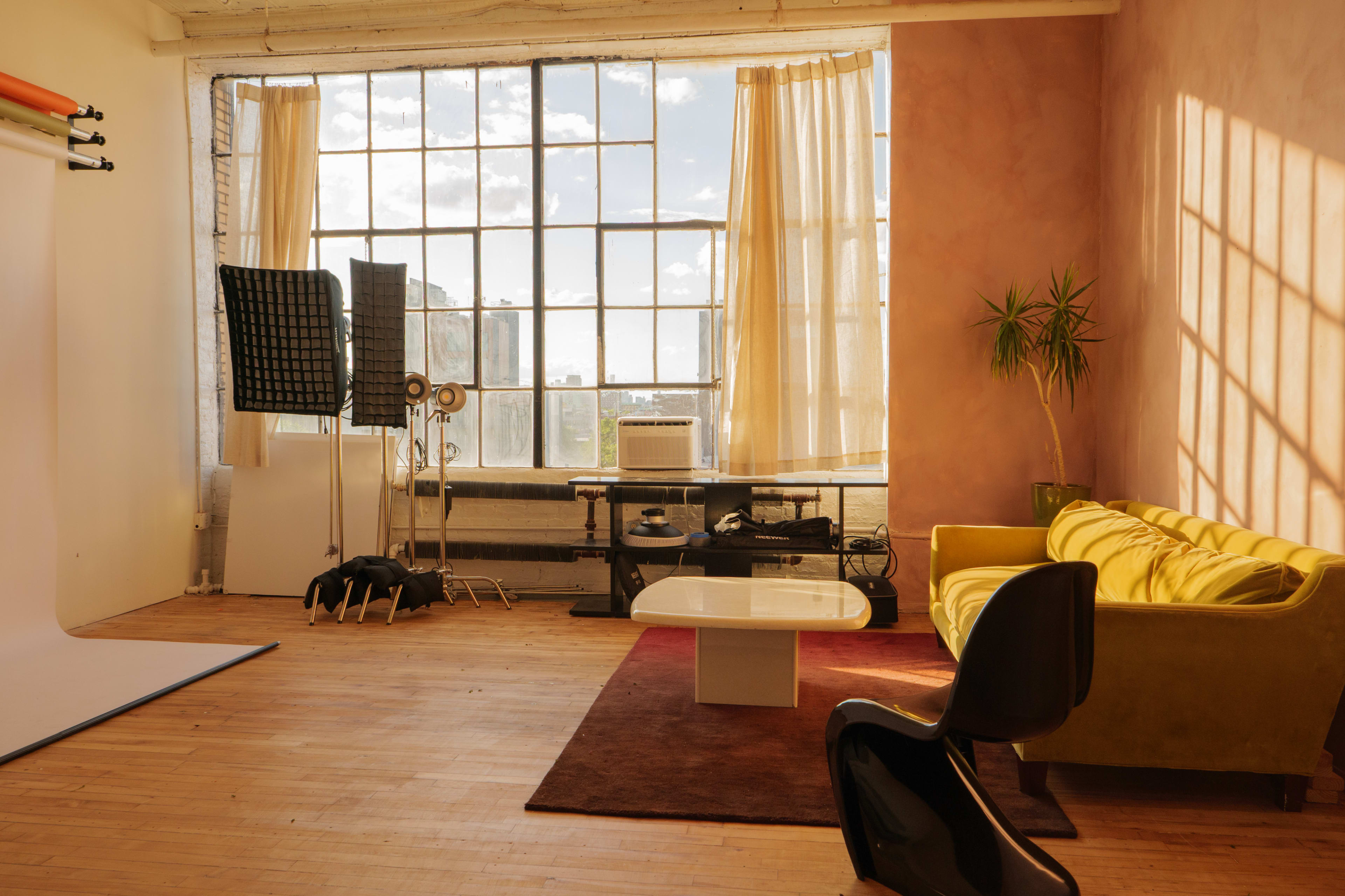The image shows a spacious interior studio with large windows, a yellow couch, a coffee table, and various lighting equipment set up against a wall.