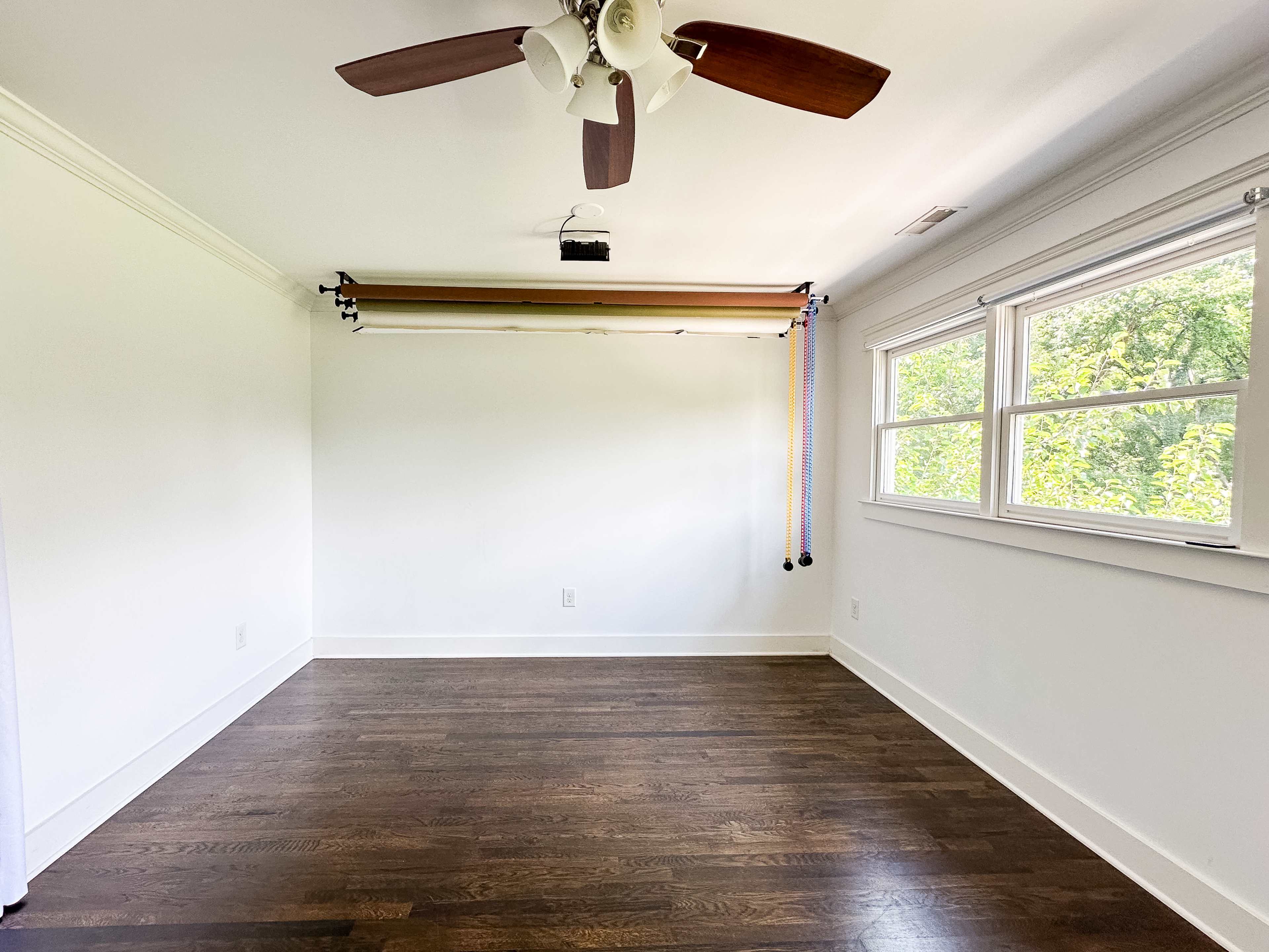 The image shows a bright, empty room with a wooden floor, a ceiling fan, and a window overlooking greenery.