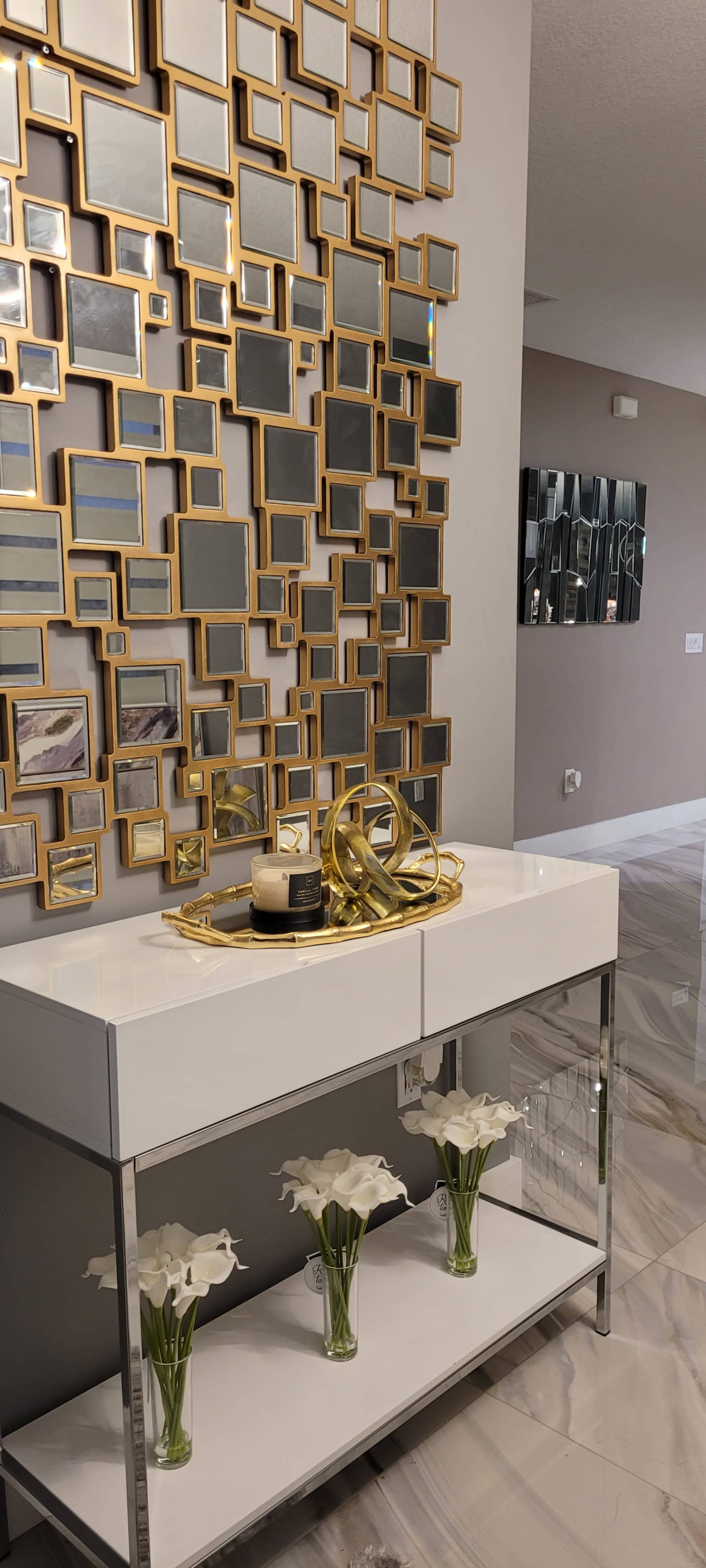 A modern entryway features a white console table adorned with flowers and decorative items, set against a wall of small, gold-framed mirrors.