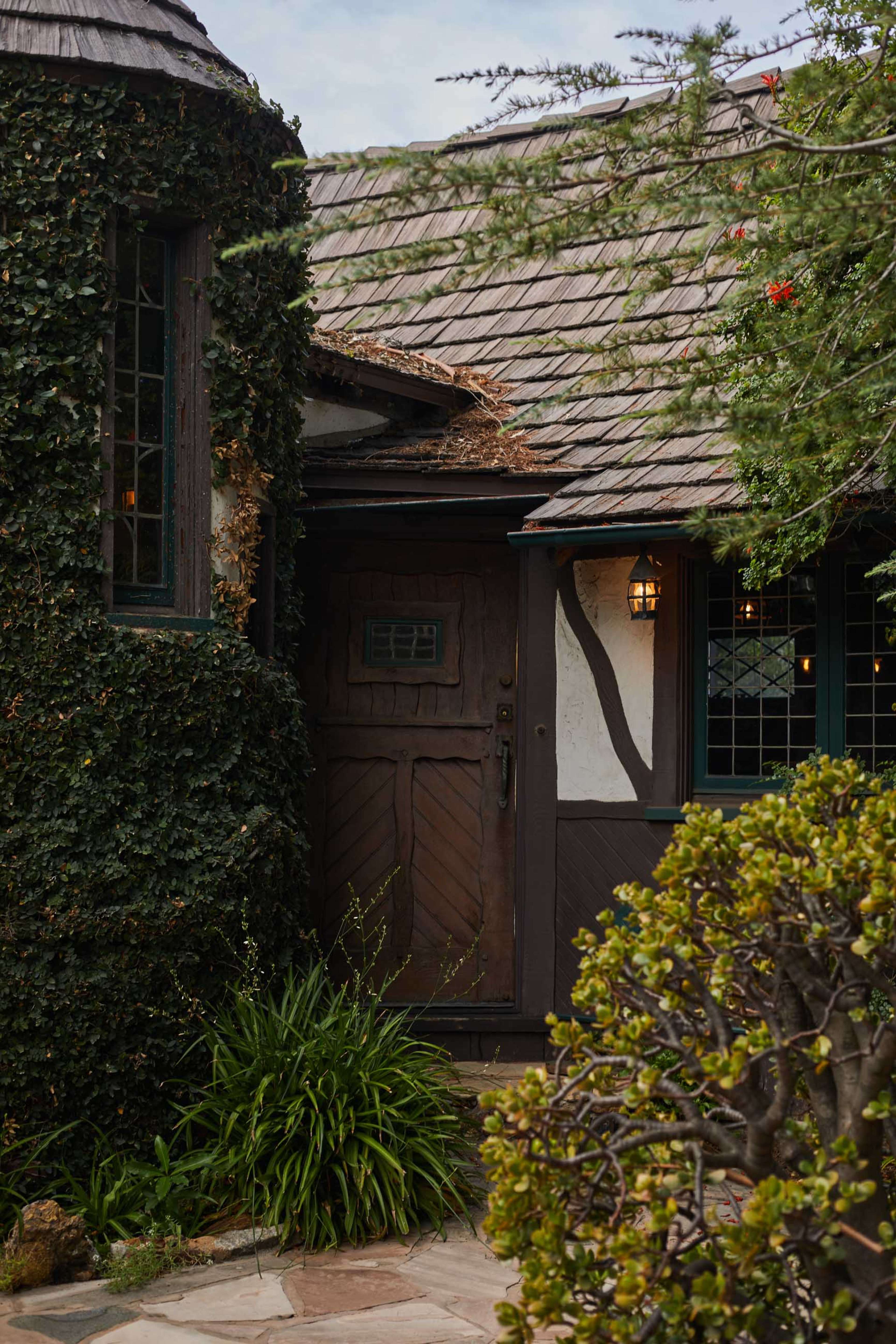 A rustic house with ivy-covered walls features a wooden door and a stone pathway lined with low shrubs.