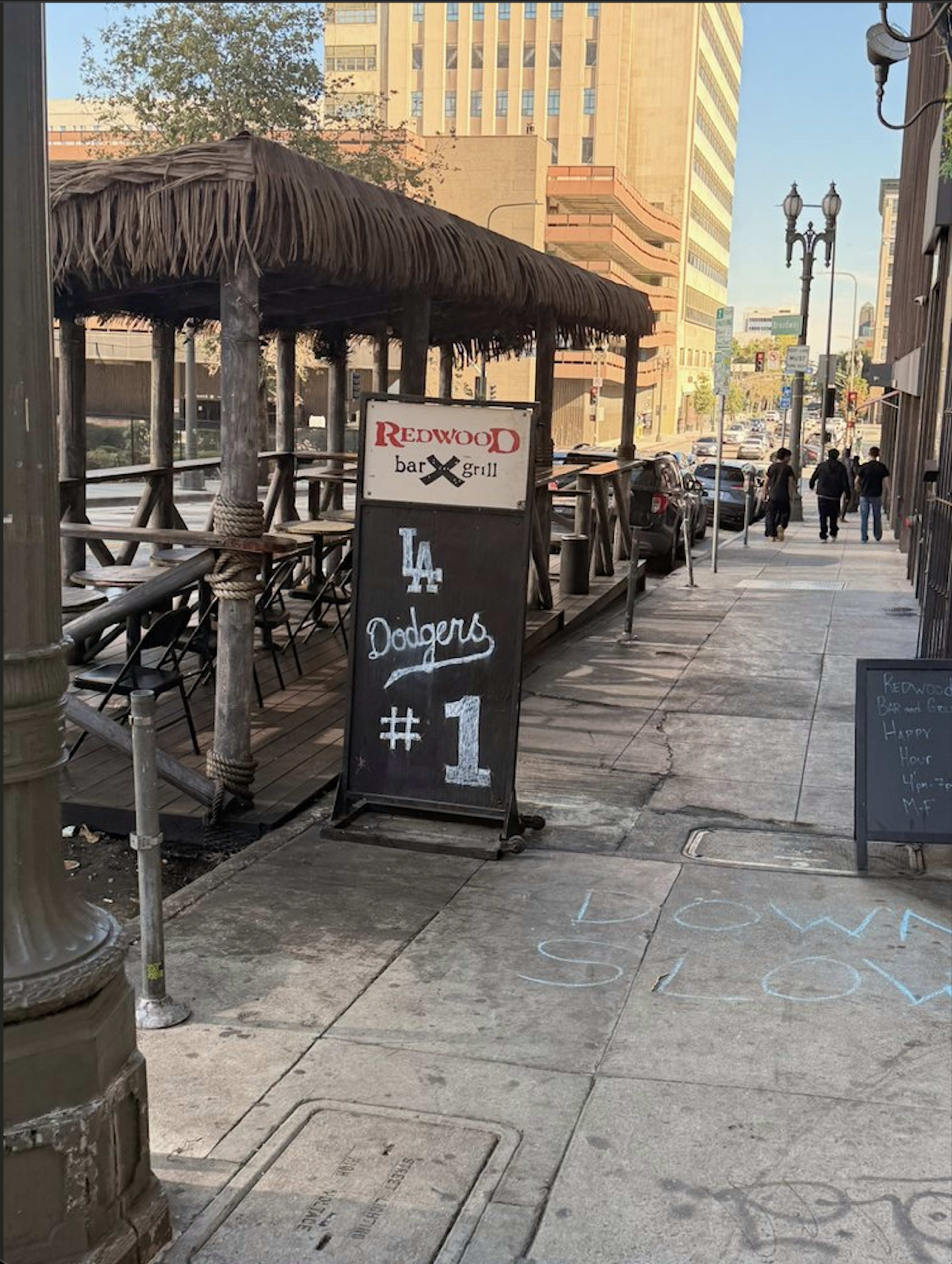 The image shows a sidewalk in an urban area with a thatched roof patio outside a restaurant, featuring a sign that promotes the Los Angeles Dodgers.