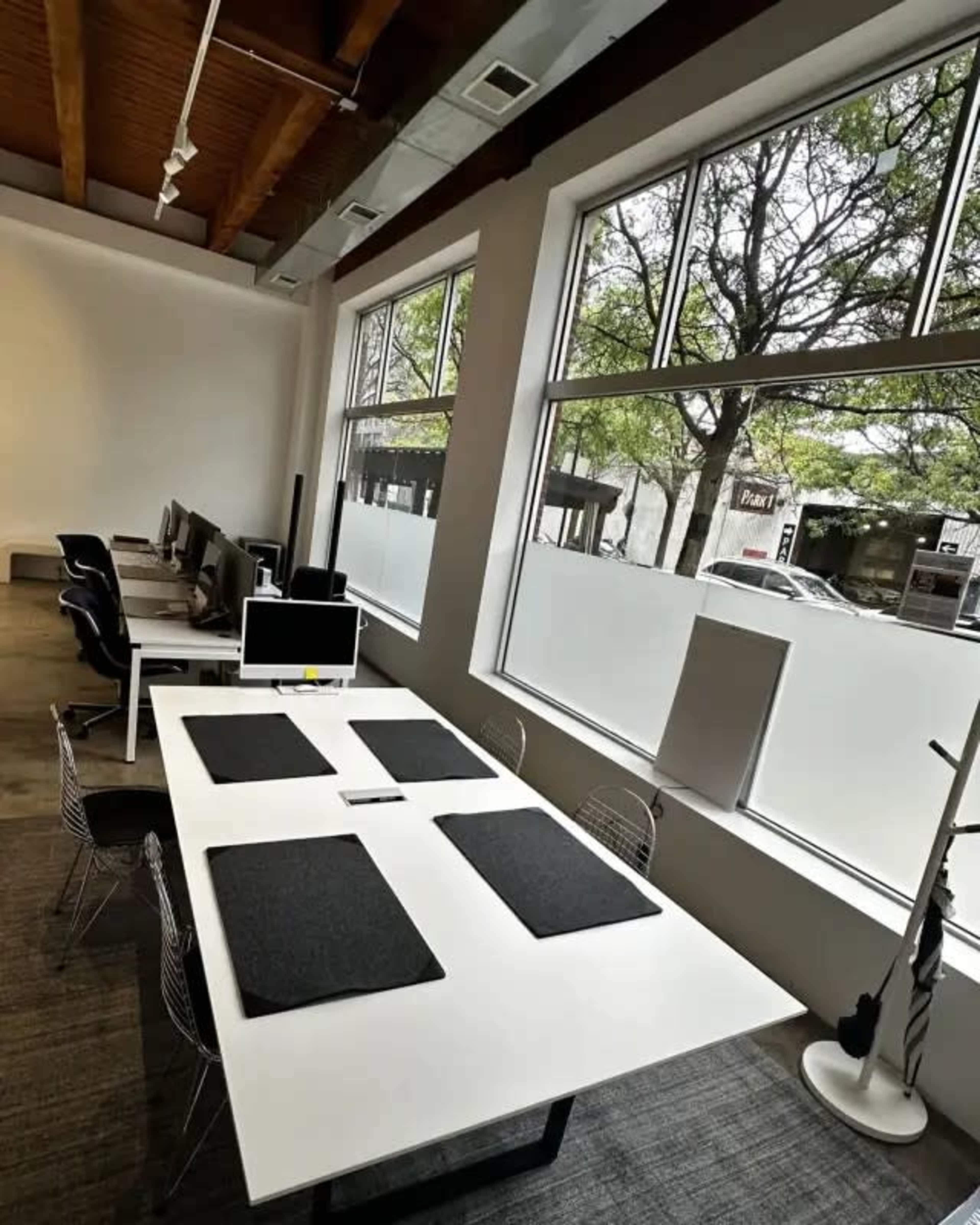 The image shows a modern office space featuring a long white table surrounded by chairs, with large windows letting in natural light.