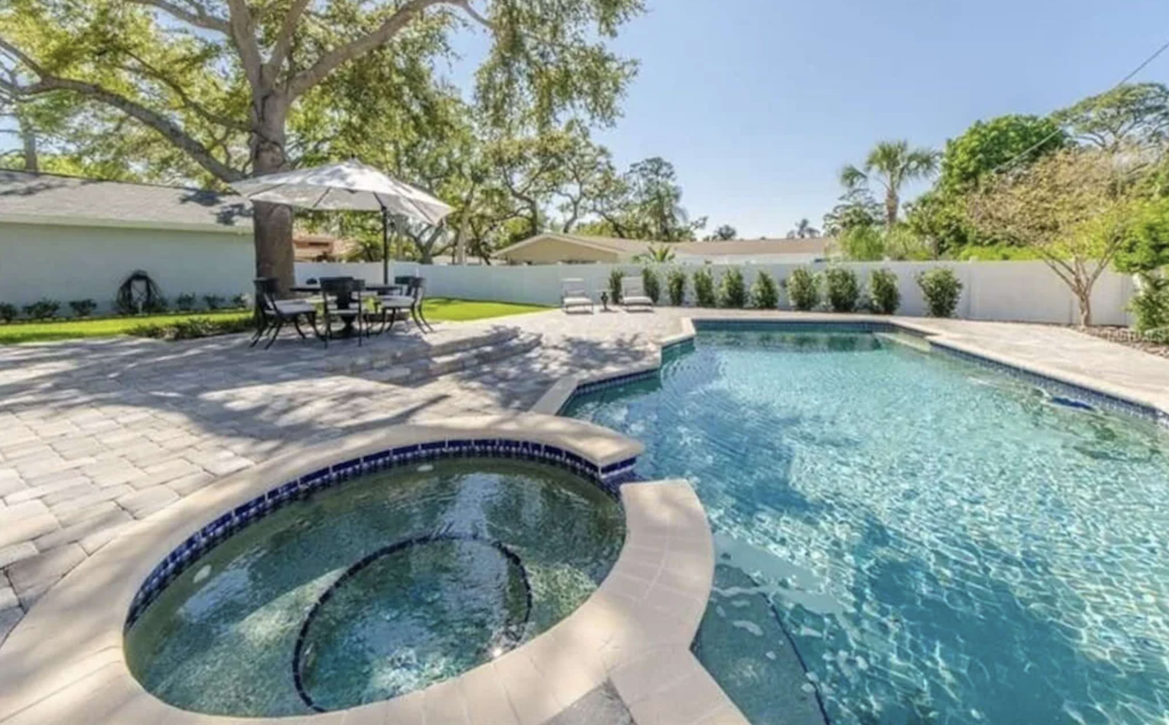 A swimming pool with a hot tub, surrounded by paved stones and lawn furniture under an umbrella in a residential backyard.