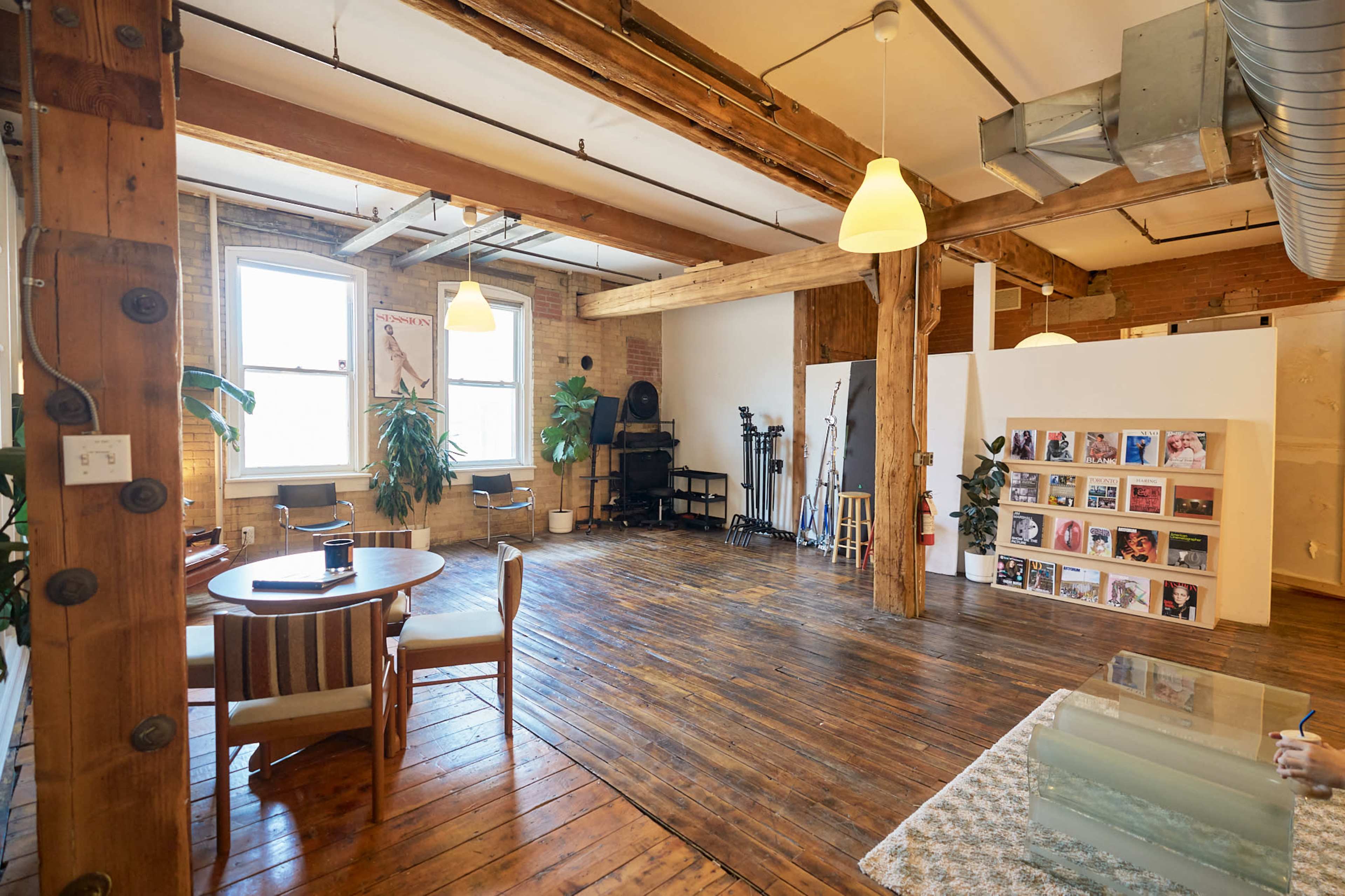 The image shows a spacious, loft-style room with wooden floors, exposed beams, and a seating area featuring a round table and chairs, alongside a wall displaying framed photographs and equipment for creative work.