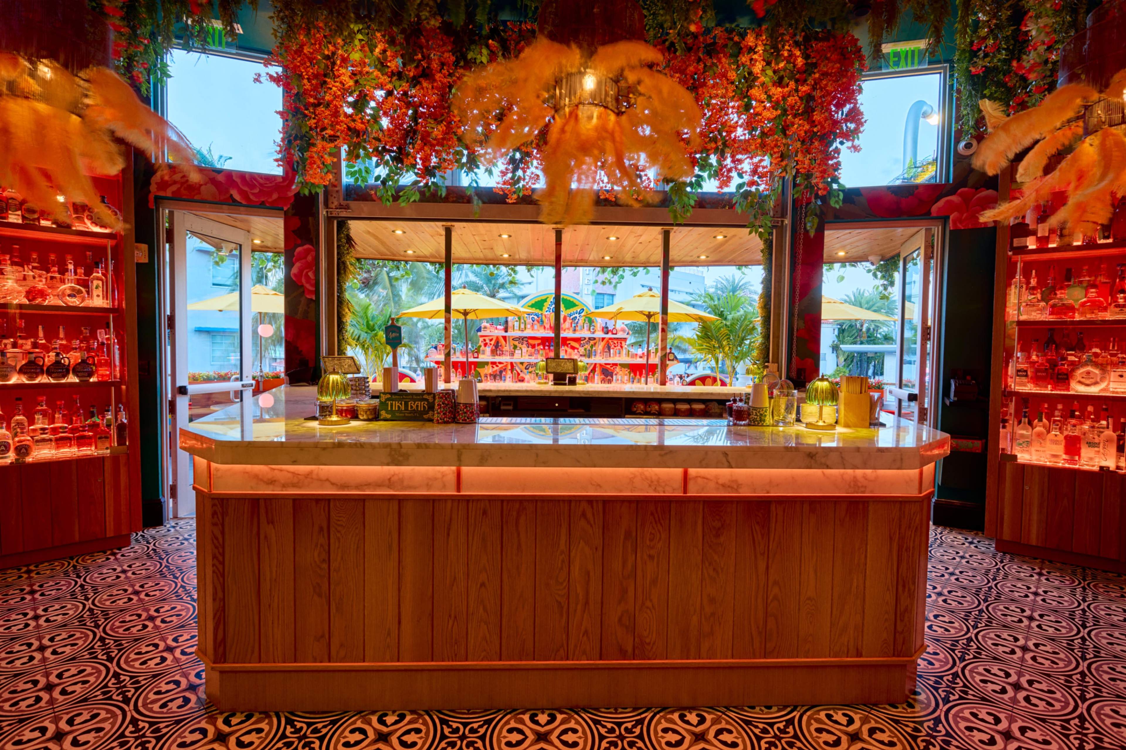 The image shows a vibrant bar area adorned with floral decorations, featuring a marble counter and illuminated shelves filled with various bottles, overlooking a patio with yellow umbrellas.