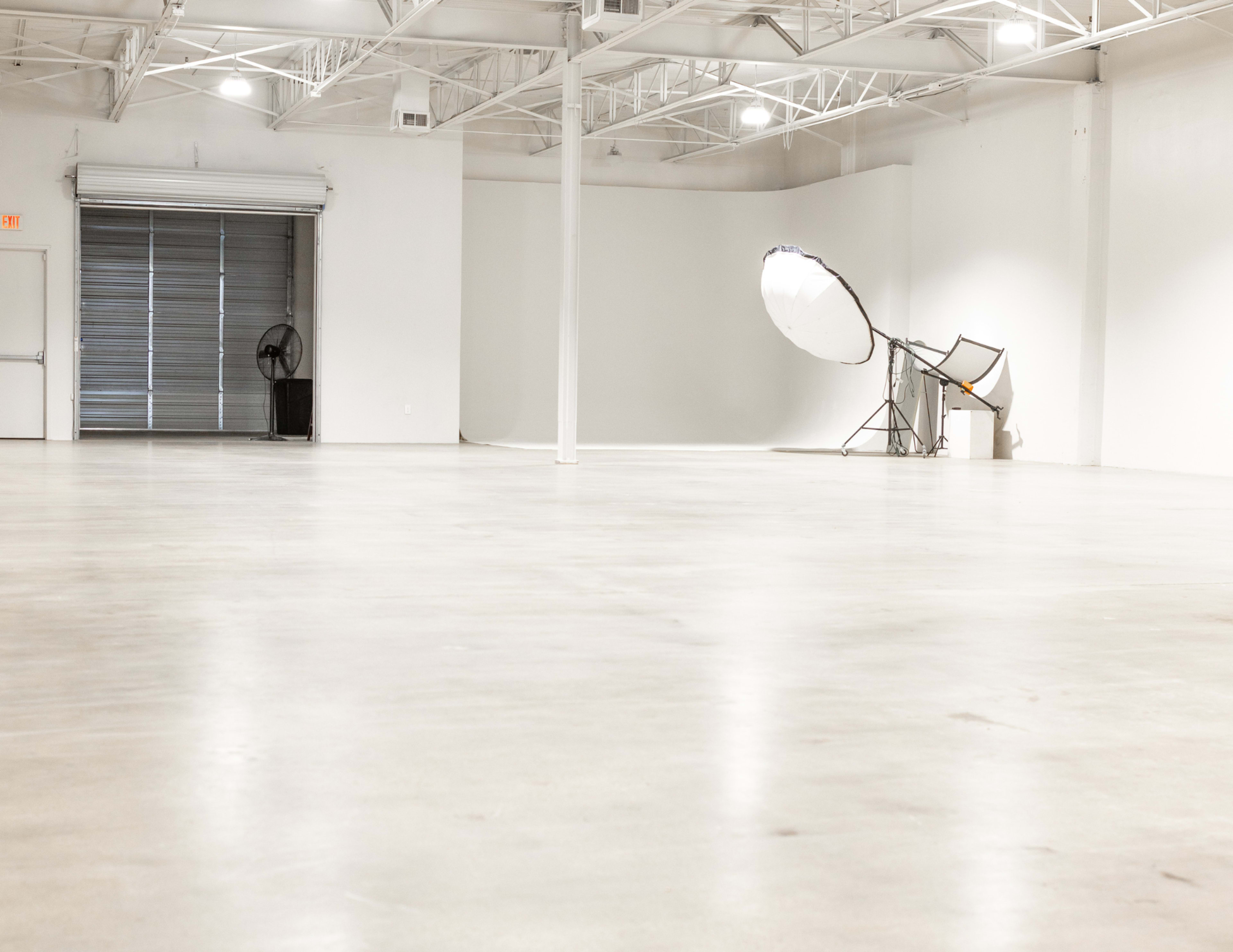 A spacious, empty studio with a concrete floor, large softbox lighting setup, and a fan near a closed roll-up door.