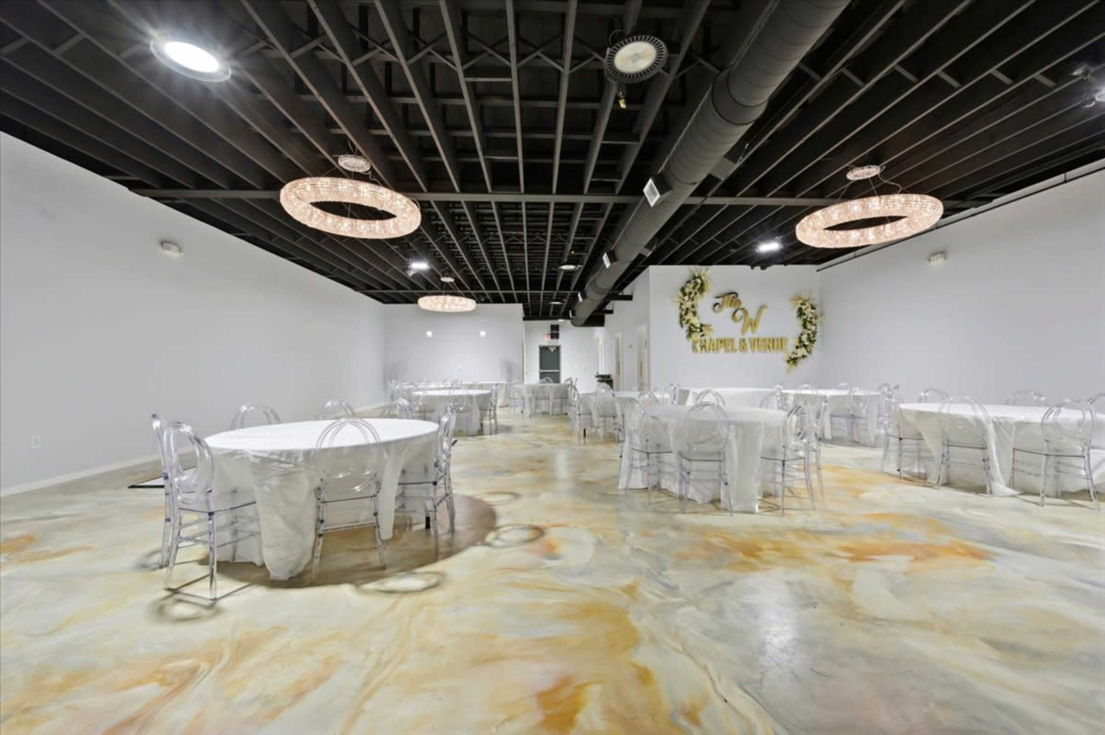 The image shows a spacious event venue featuring several round tables covered with white tablecloths, clear chairs, and modern lighting fixtures on a polished floor.