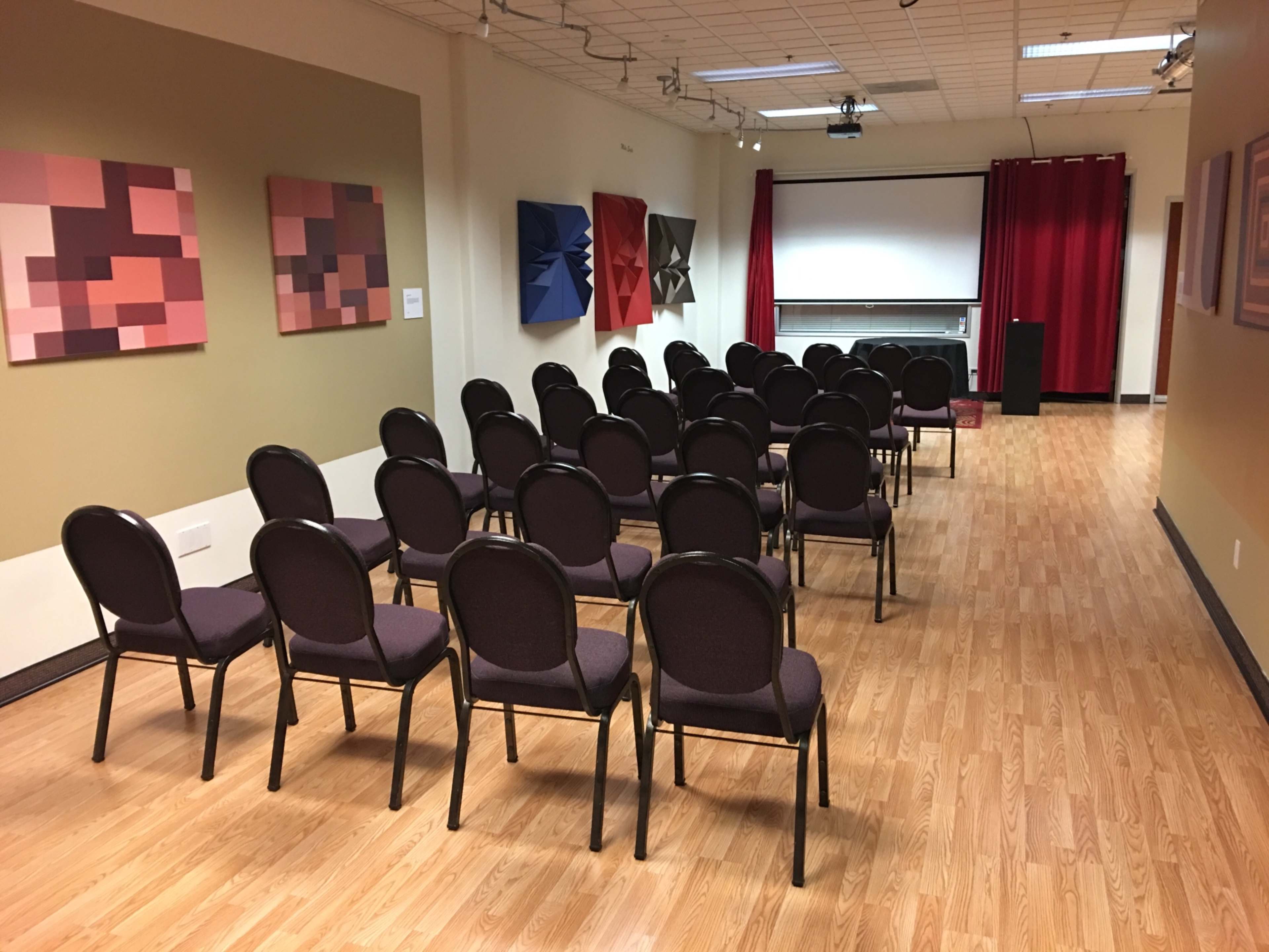 A modestly arranged room featuring rows of chairs facing a screen, with abstract artwork on the walls and red curtains.