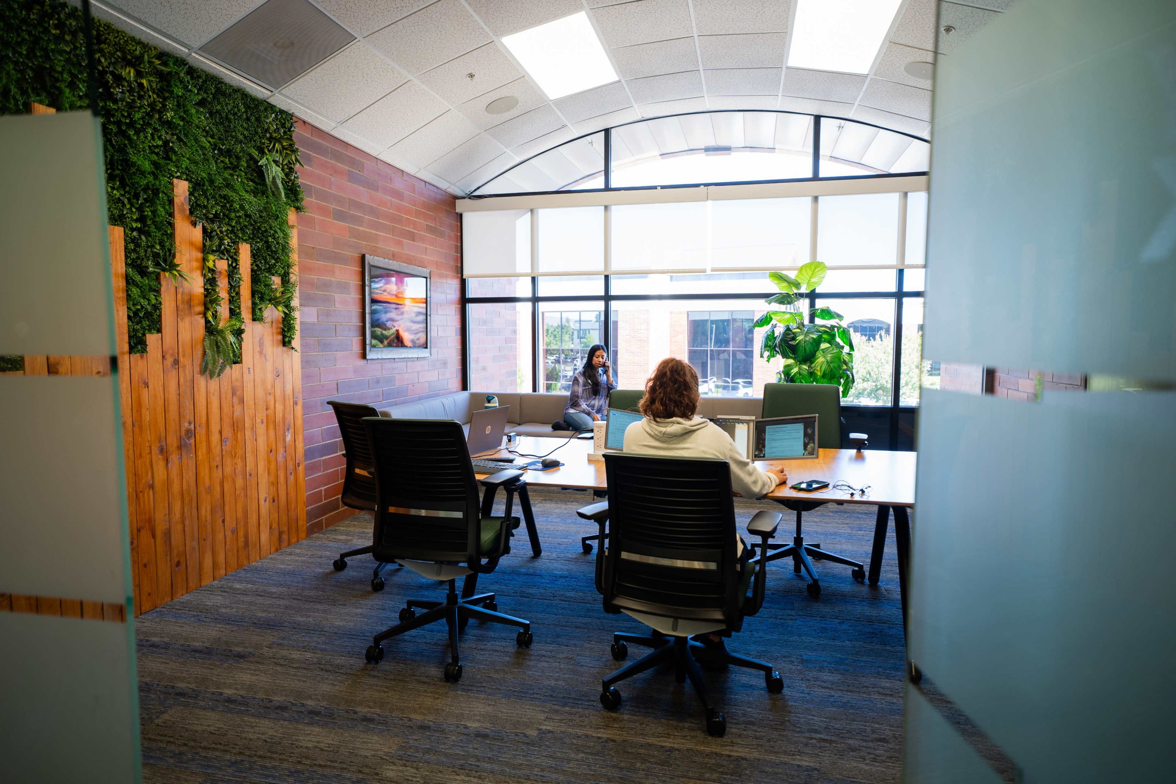 A modern office features a large conference table surrounded by chairs, with two individuals working on laptops near a plant and a window.
