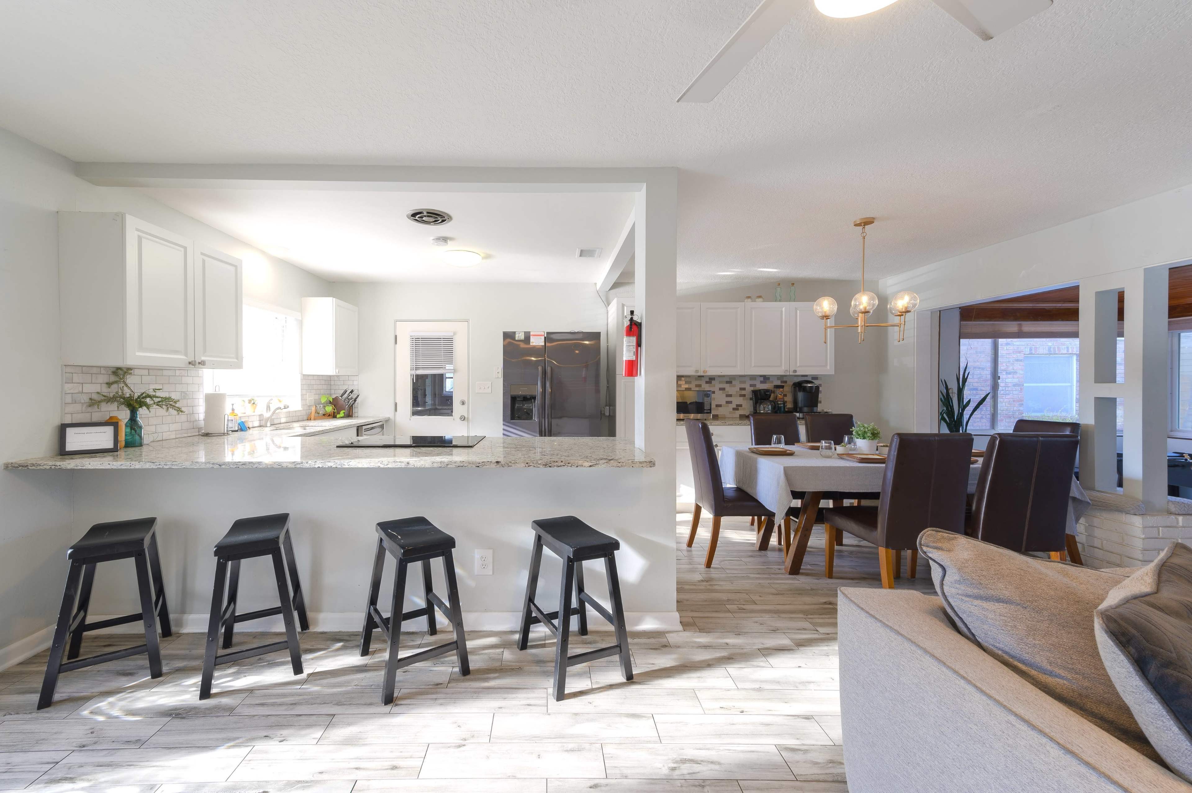 The image shows a modern kitchen and dining area with a granite countertop and black bar stools, featuring a dining table set for a meal.