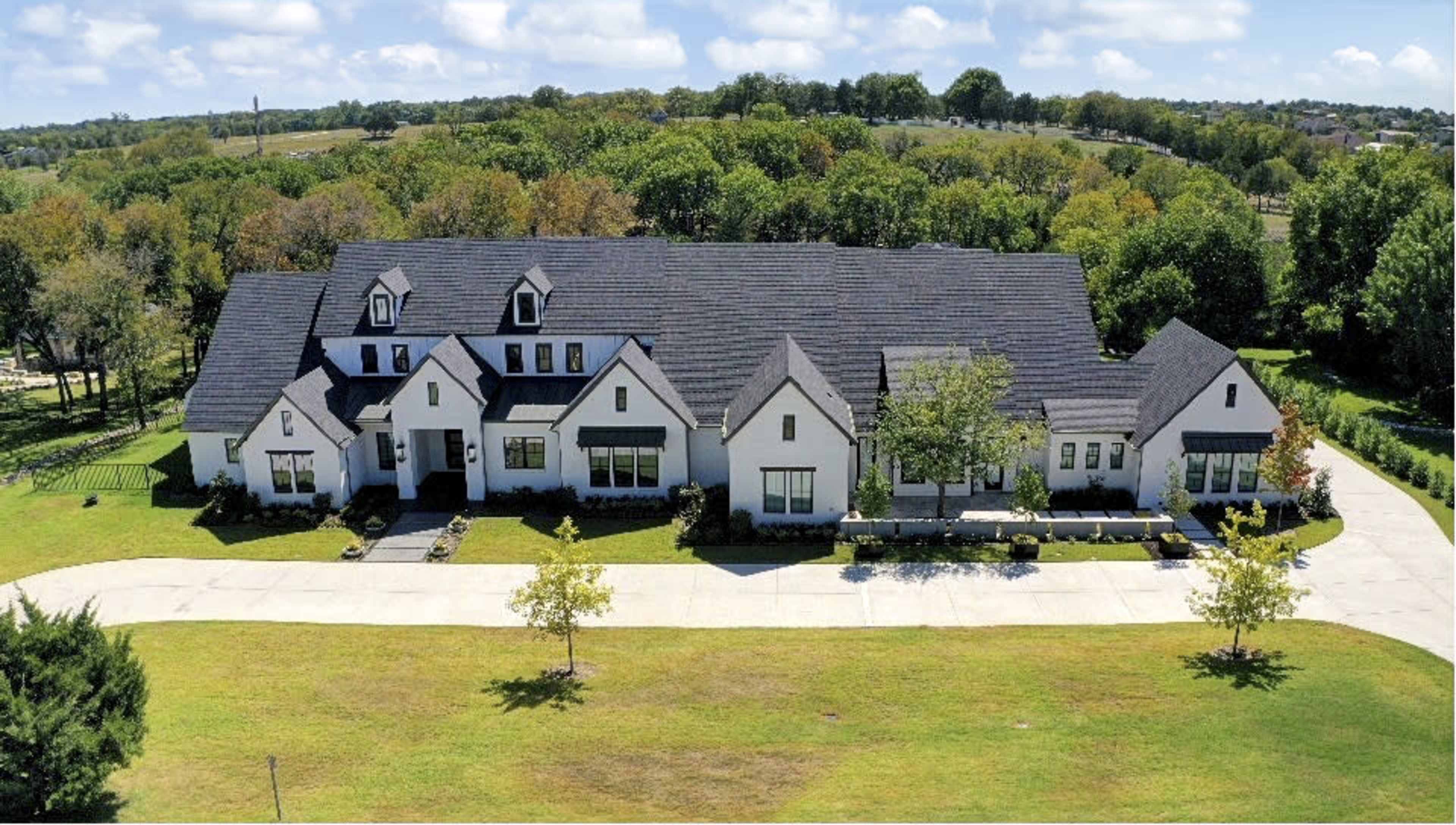 A large, modern farmhouse-style house with multiple gabled roofs, situated on a spacious green lawn with scattered trees in the background.