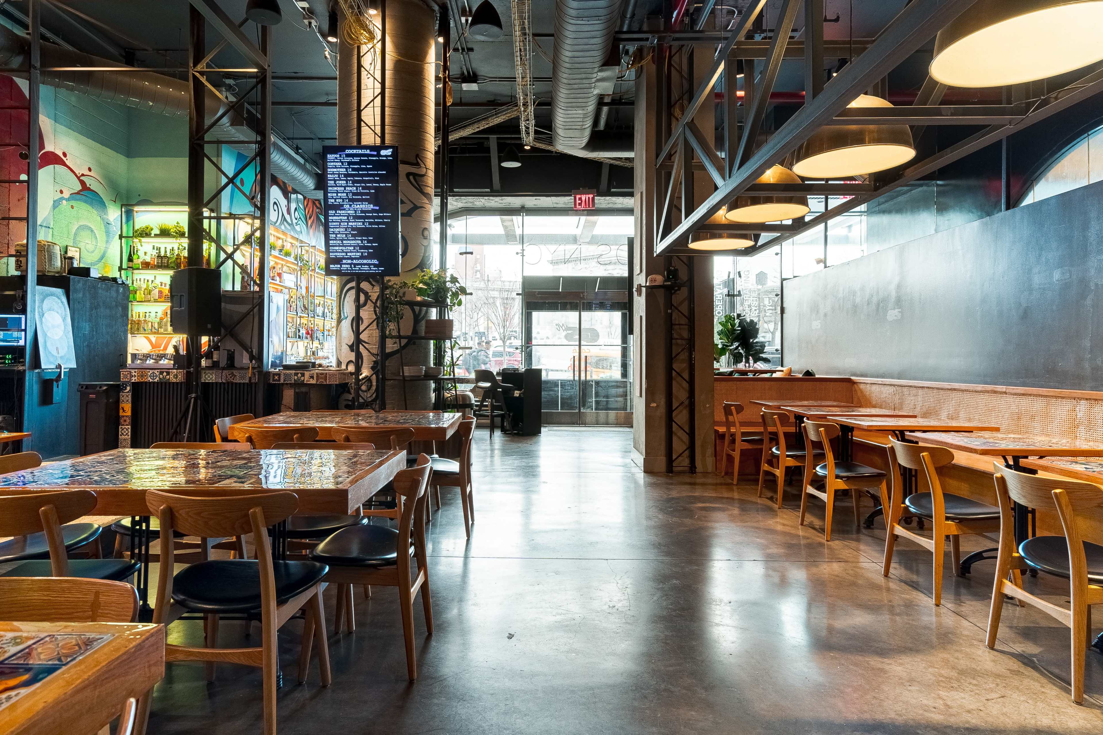 An industrial-style restaurant interior with wooden tables, metal accents, and large windows providing natural light.
