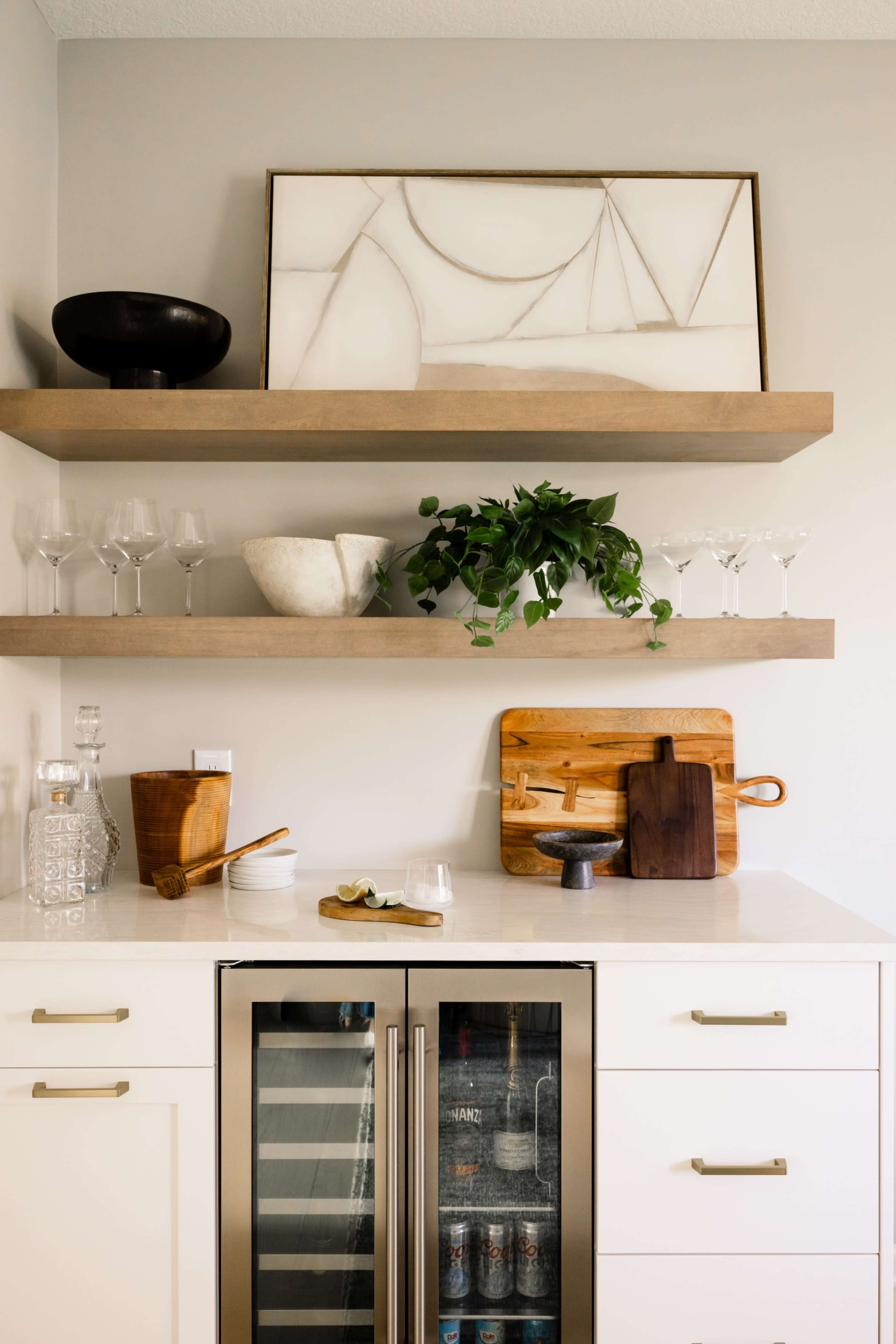The image shows a modern kitchen with wooden shelves holding glassware and decorative items above a bar area featuring a wine fridge and wooden cutting boards.
