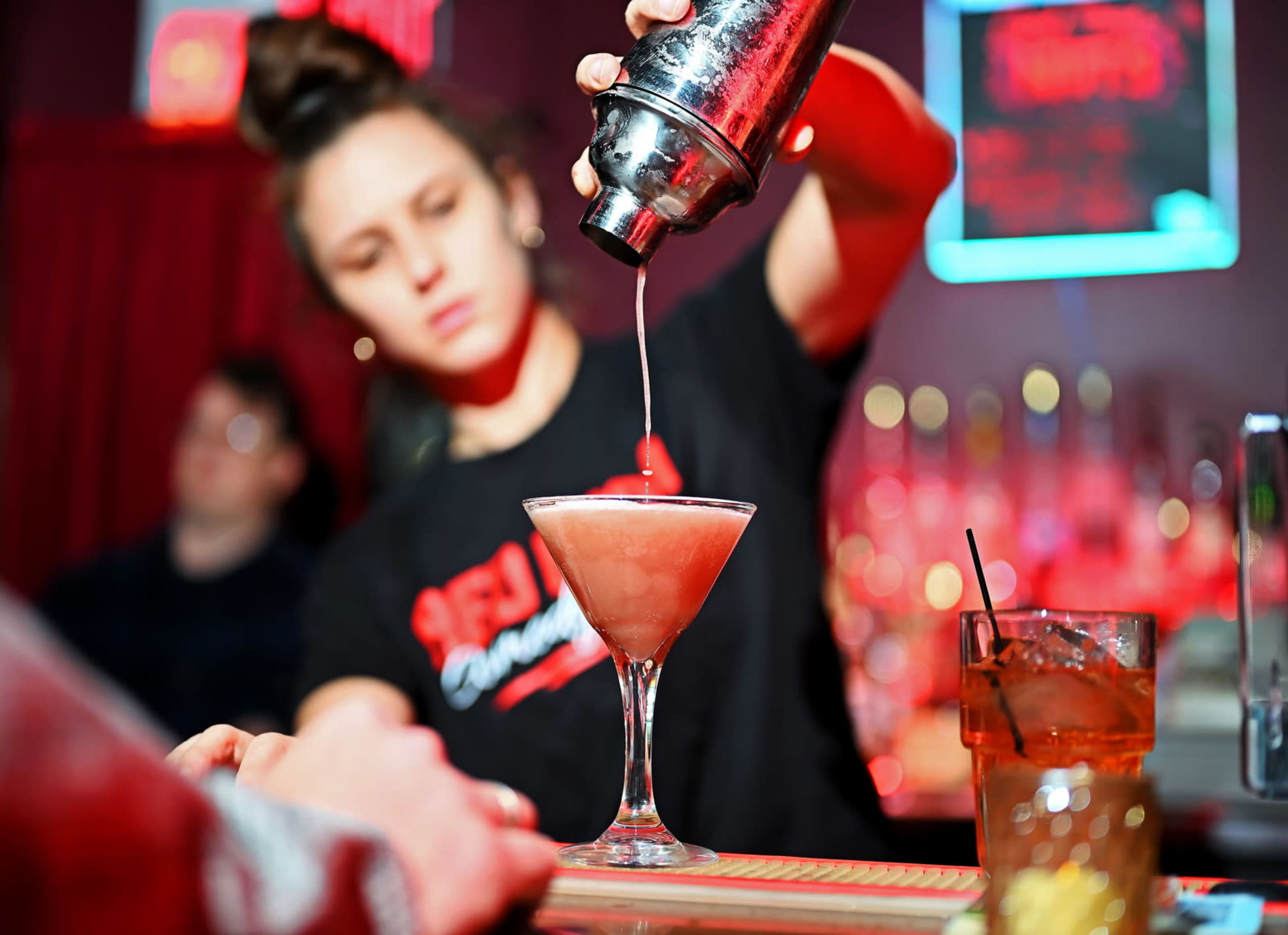 A bartender pours a pink cocktail from a shaker into a glass at a vibrant bar scene.
