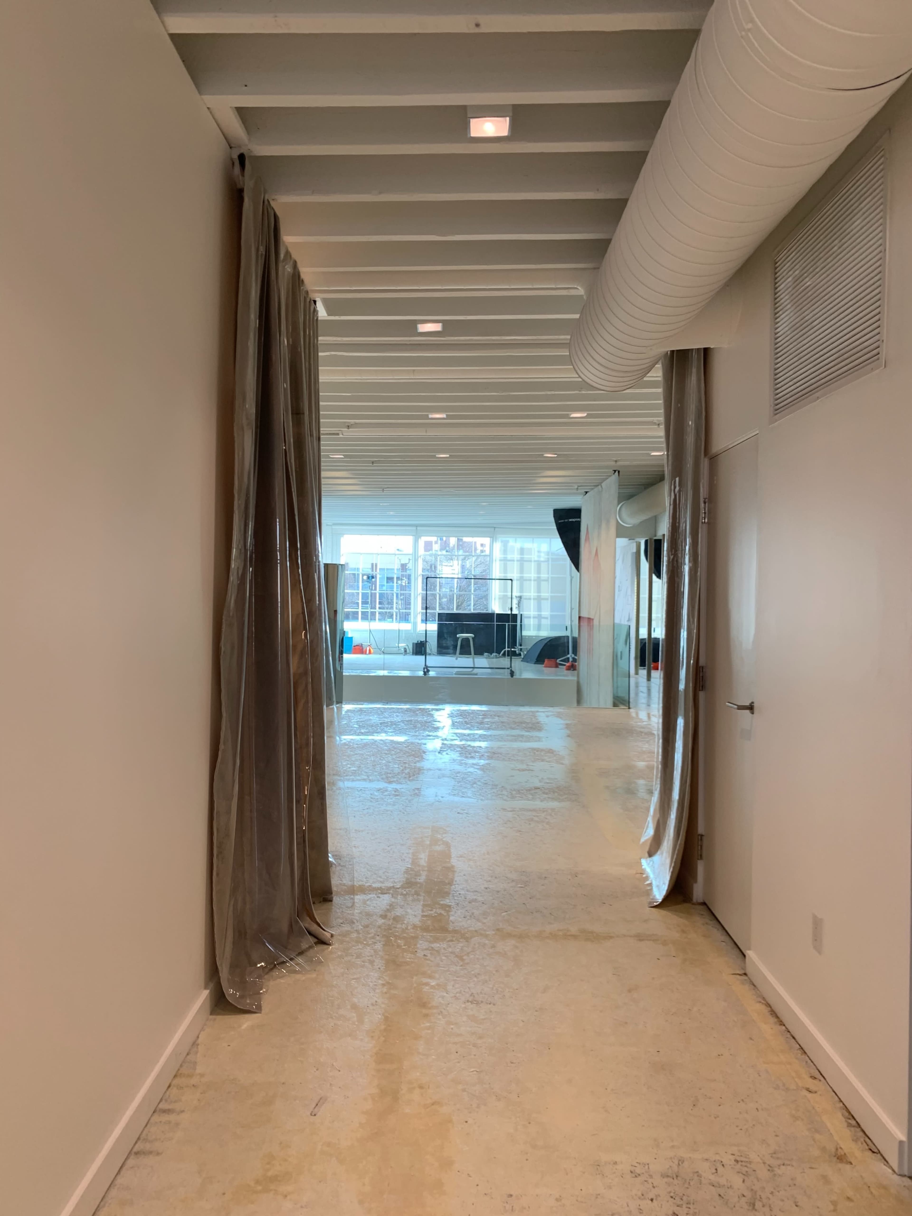 The image shows a long, narrow hallway with a section of translucent curtains on one side and a view of a bright room at the end, featuring large windows and visible furniture.