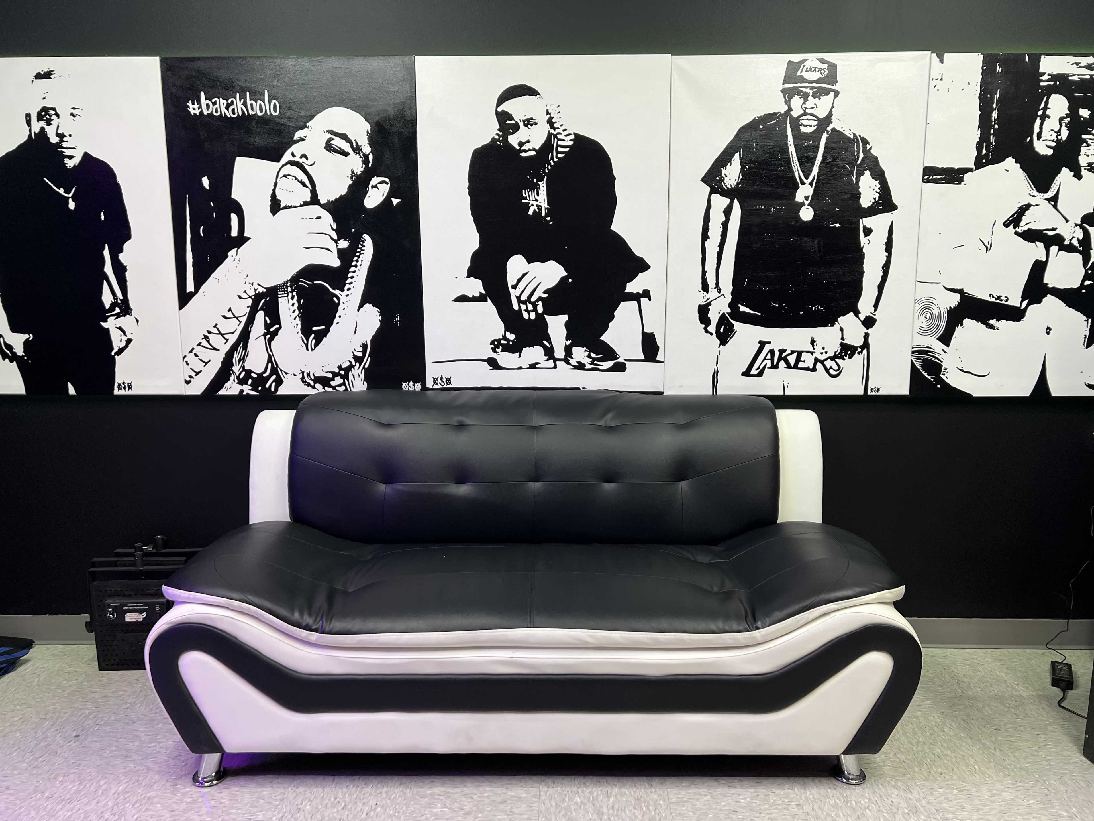 The image shows a black and white couch in front of a wall featuring large black and white portraits of men.