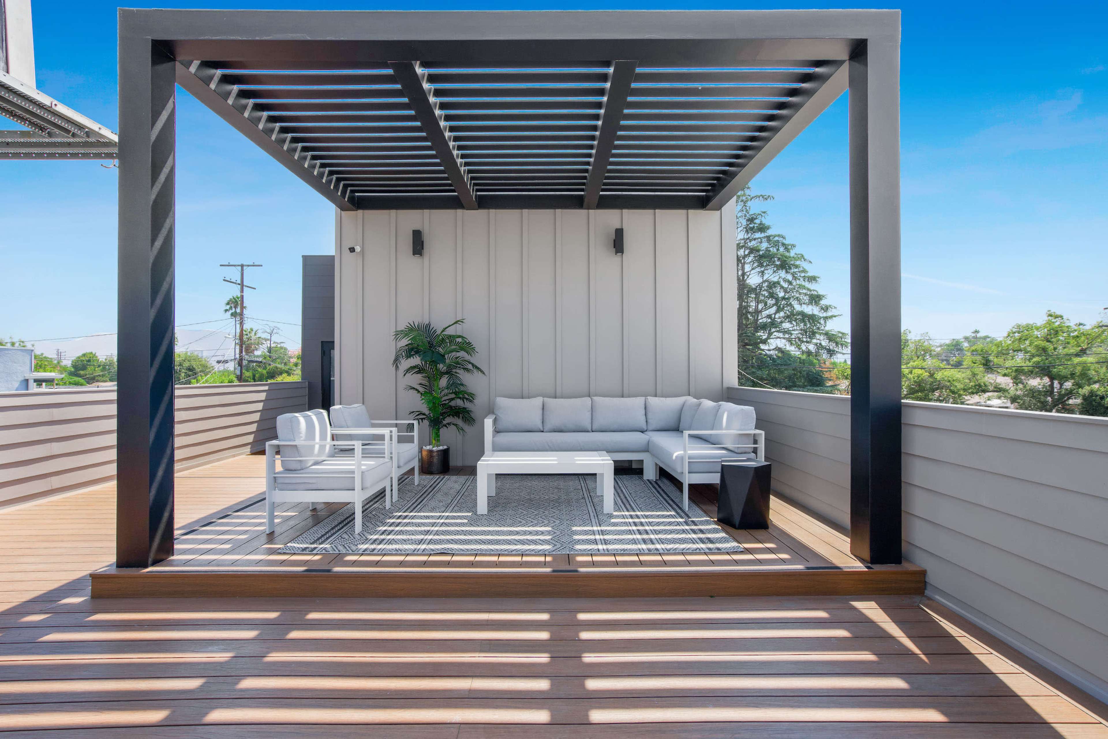 A modern outdoor seating area with a covered pergola, featuring a white sofa, two chairs, and a coffee table on a wooden deck.