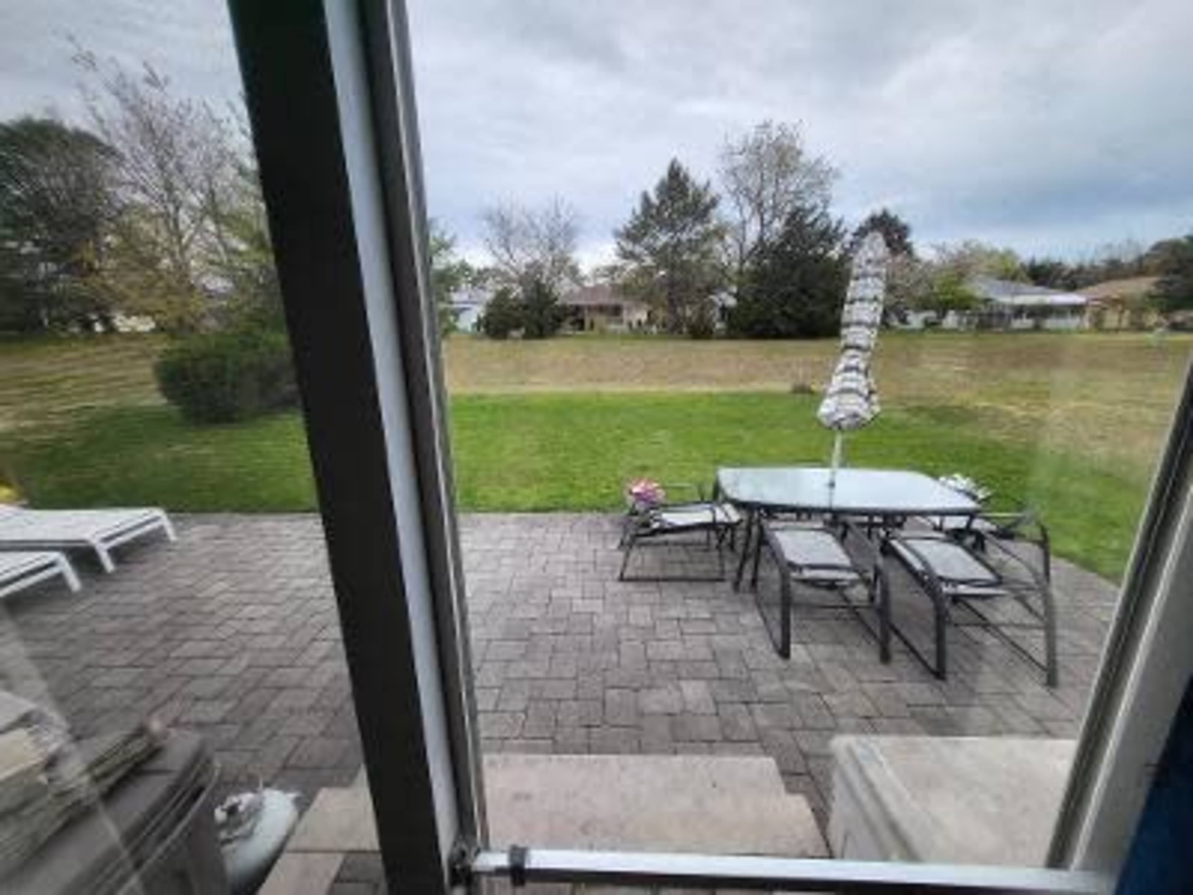 A patio area with a table and chairs under an umbrella, overlooking a grassy yard and trees beyond.