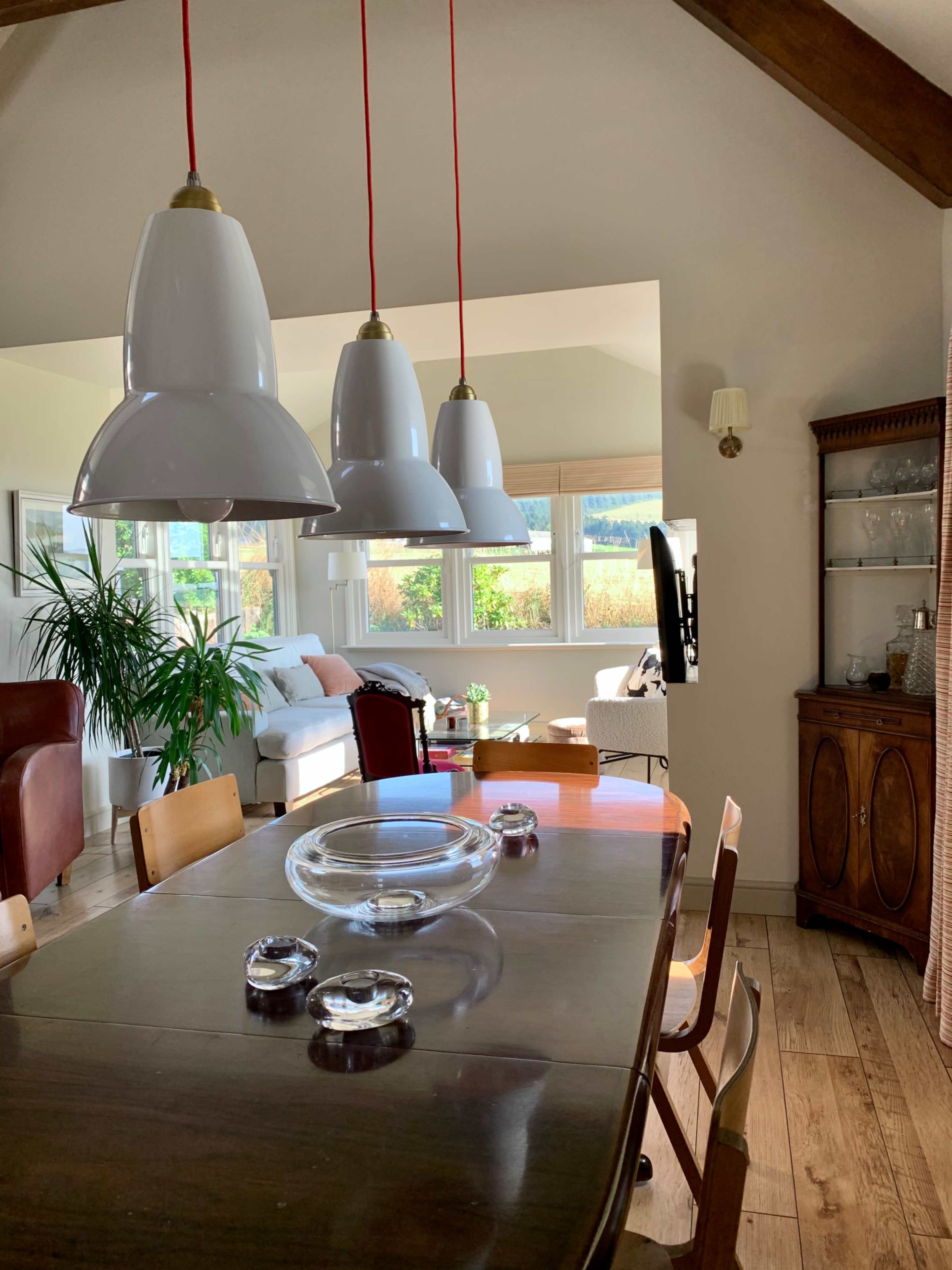 A dining table with four chairs is seen in a well-lit room featuring large windows and modern pendant lights.