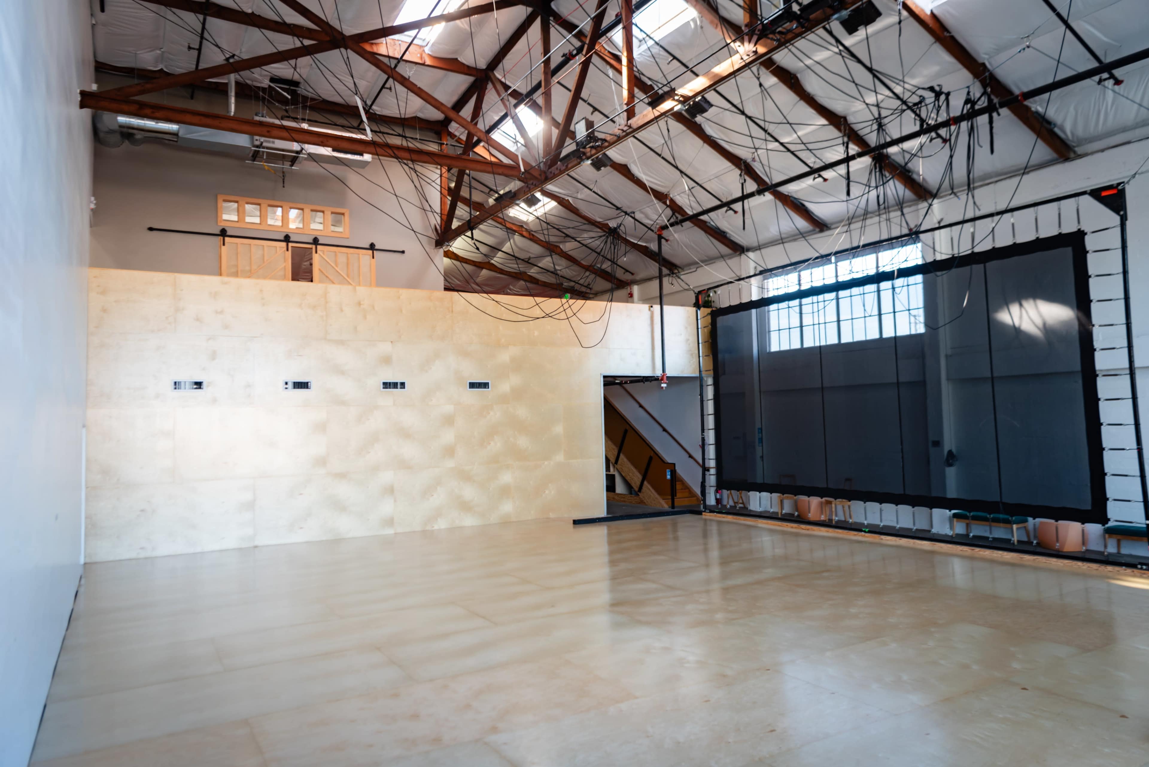 An industrial-style interior with a high ceiling, wooden beams, and a large empty wall opposite a dark projection screen.