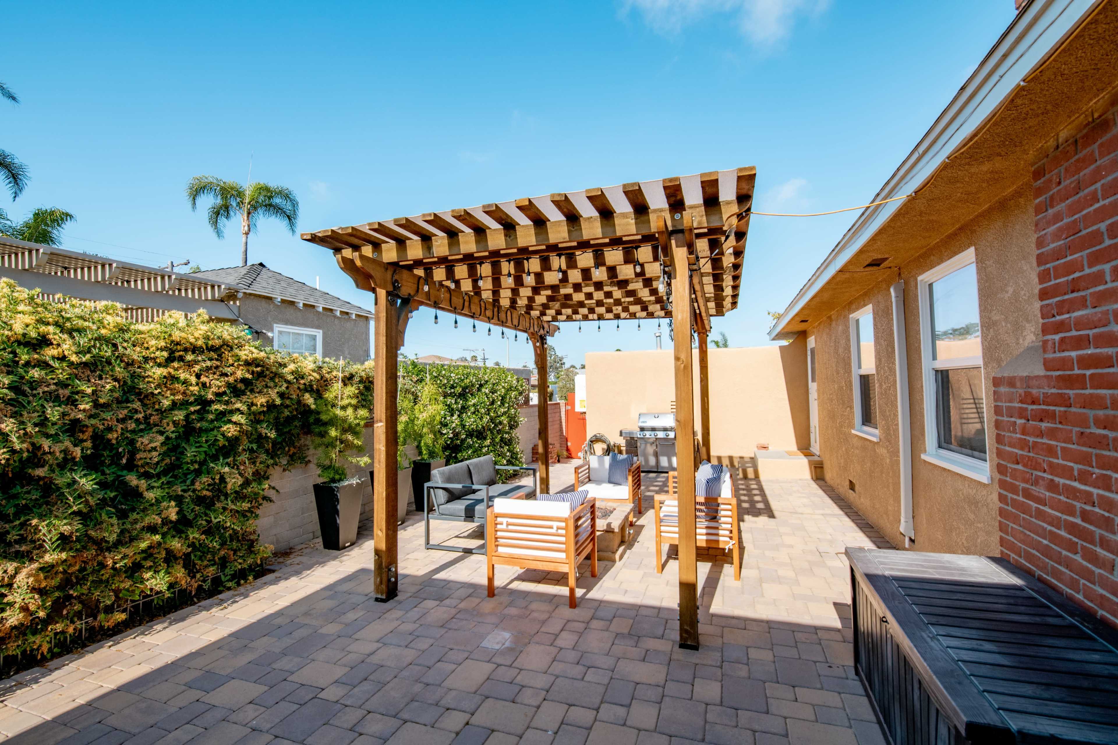 A patio area with a wooden pergola, seating arrangements, and paver stones surrounded by greenery.