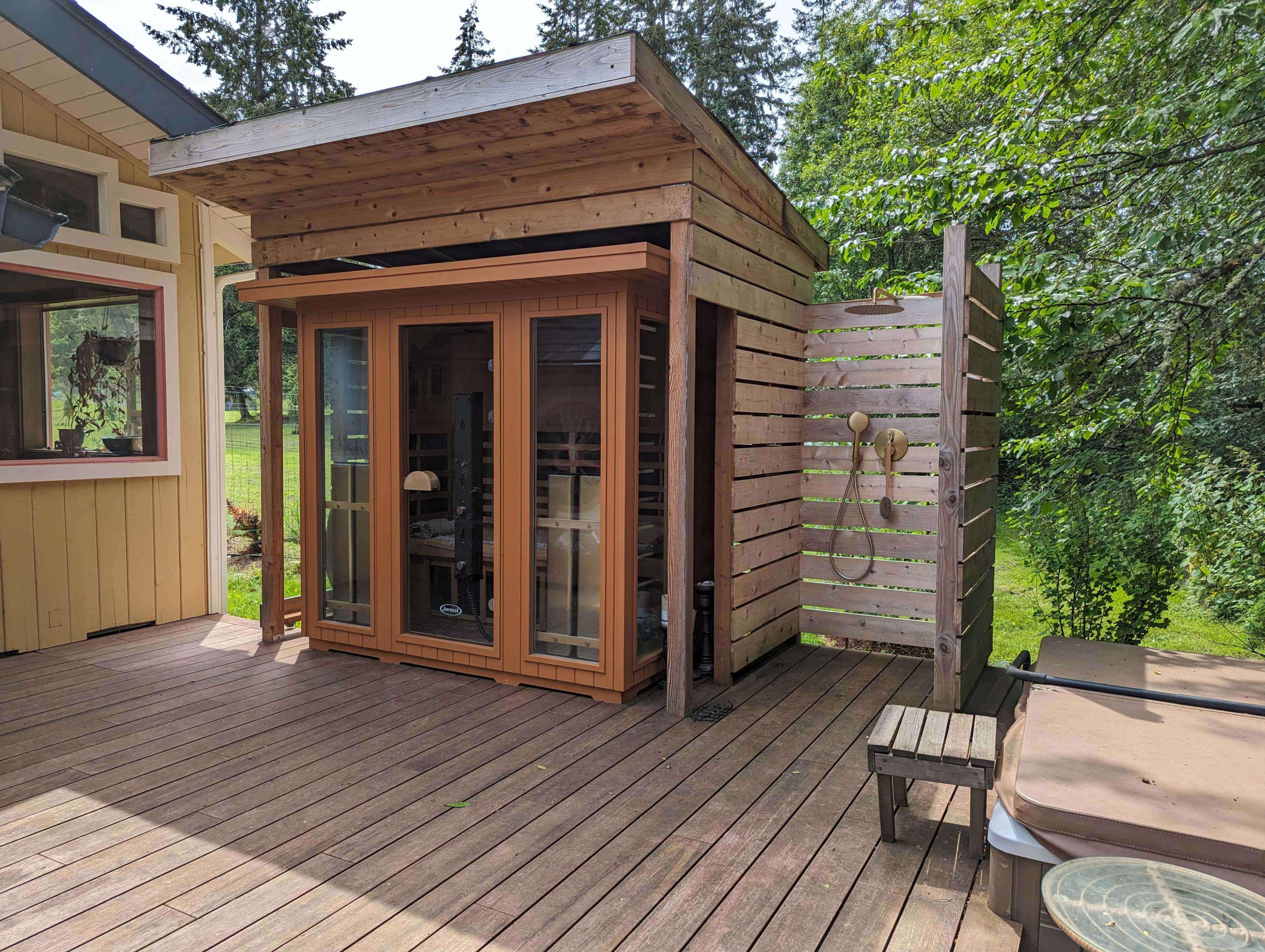 The image shows a wooden sauna with glass doors, situated on a deck surrounded by trees.