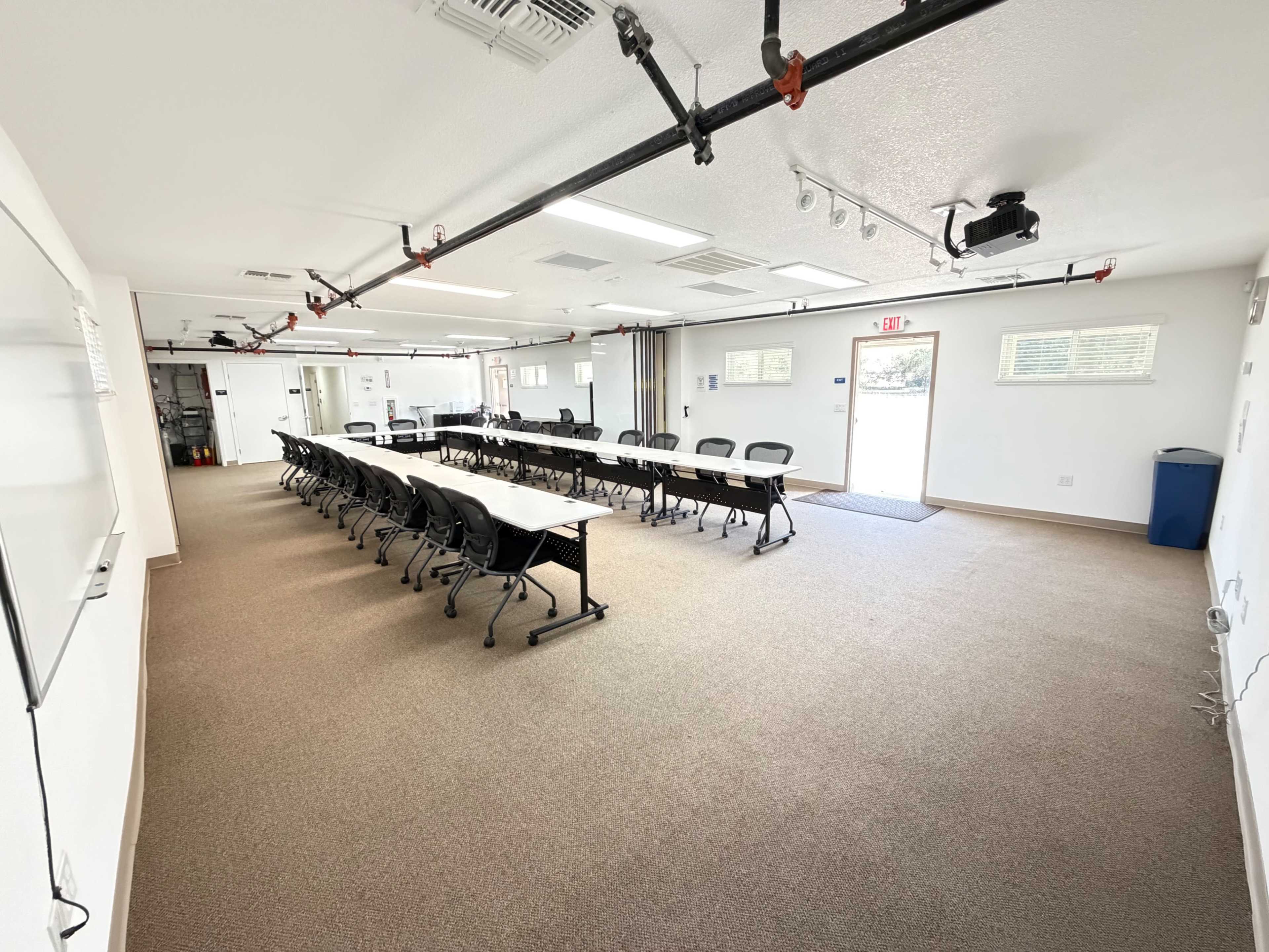 The image shows a spacious meeting room with rows of black chairs and long tables arranged facing the entrance.