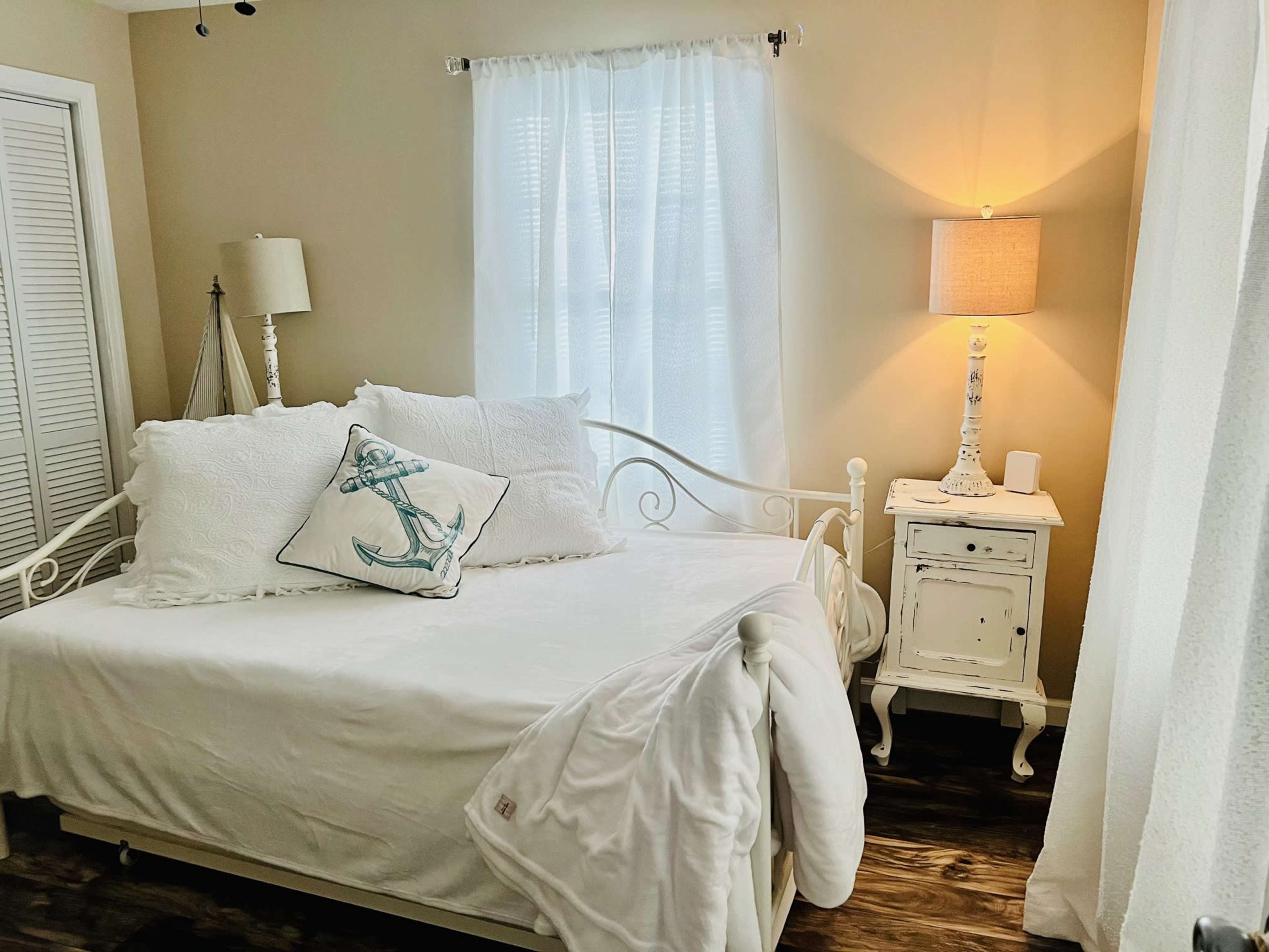 A well-lit bedroom with a white daybed, a bedside table with a lamp, and a window draped with sheer curtains.