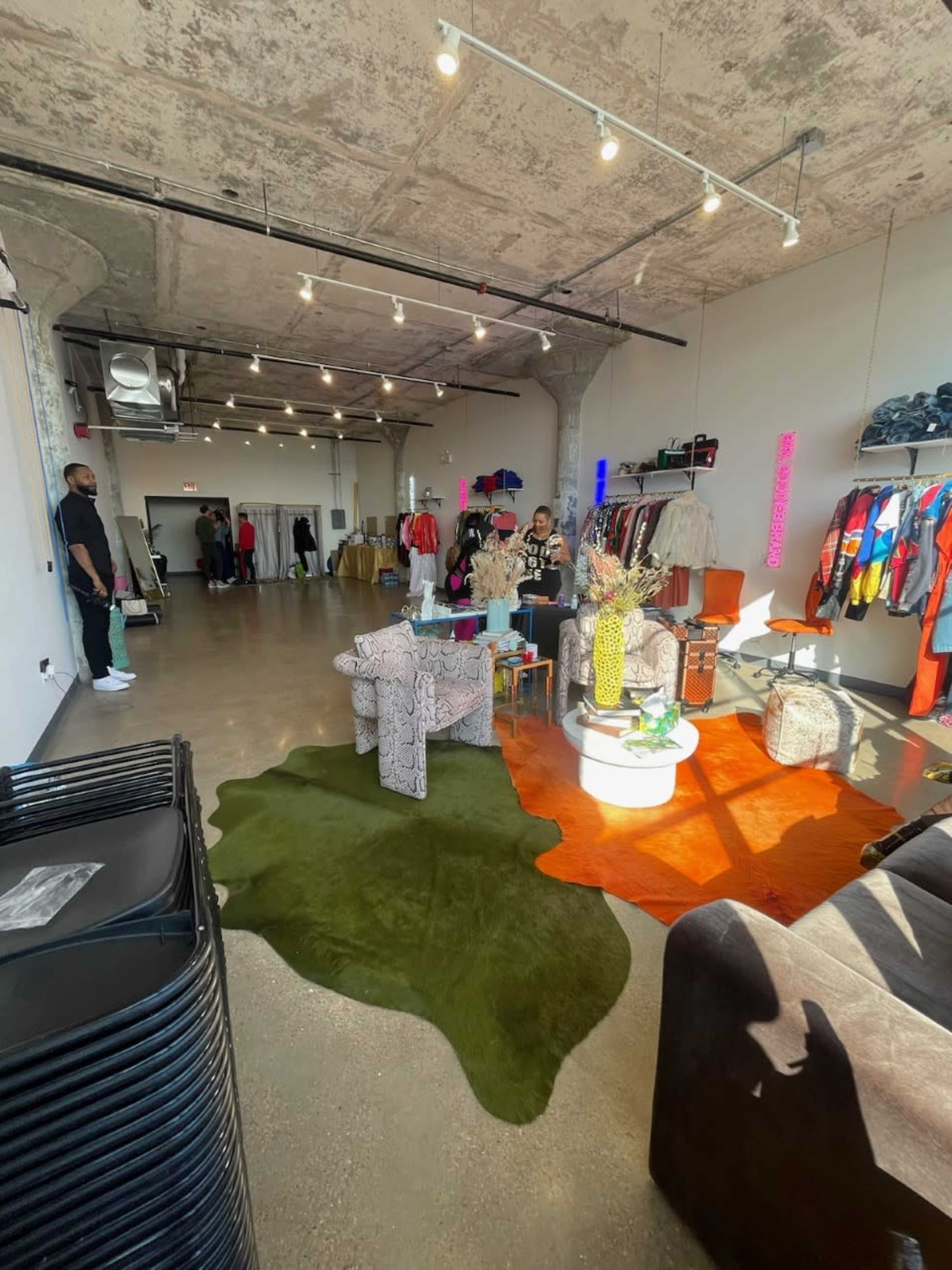 The image shows a clothing boutique with various garments displayed on racks, a seating area with colorful furniture, and a concrete ceiling.