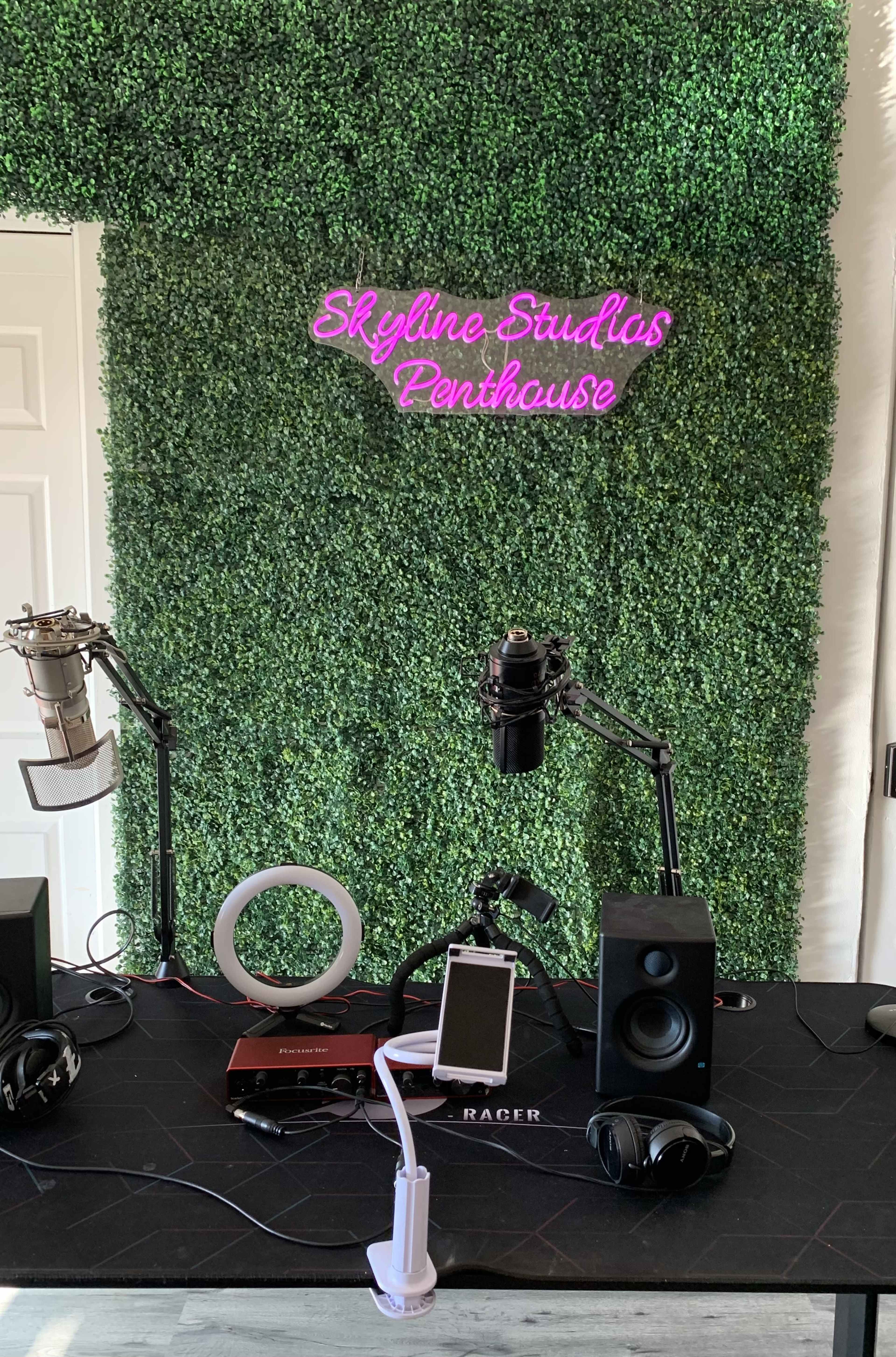 A recording studio setup with microphones, headphones, a smartphone, and speakers against a green, textured wall featuring a neon sign that reads "Skyline Studios Penthouse."