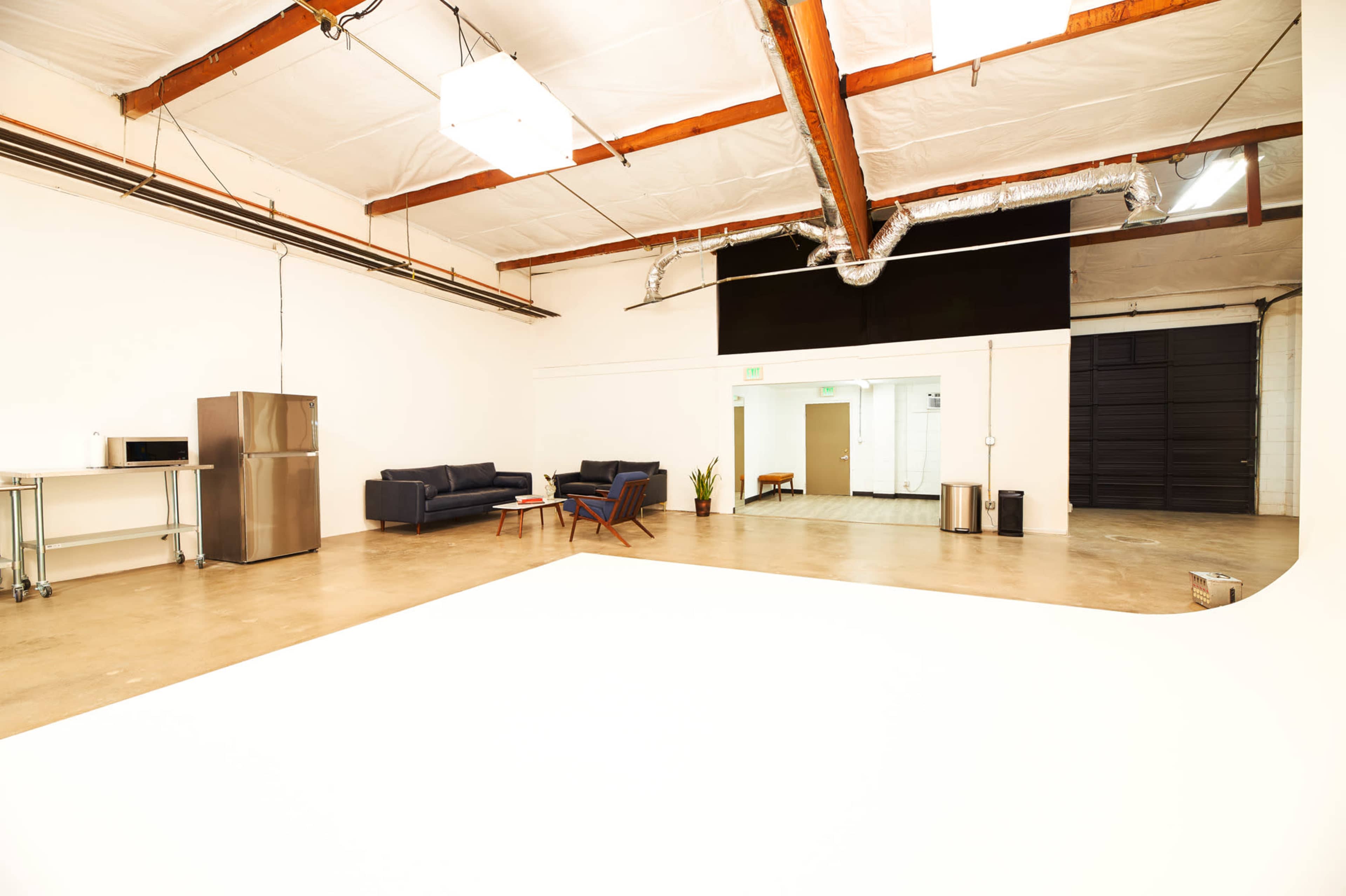 A spacious, well-lit studio features a sectional sofa, a seating area, a kitchenette with a microwave, and adjacent doors leading to another room.