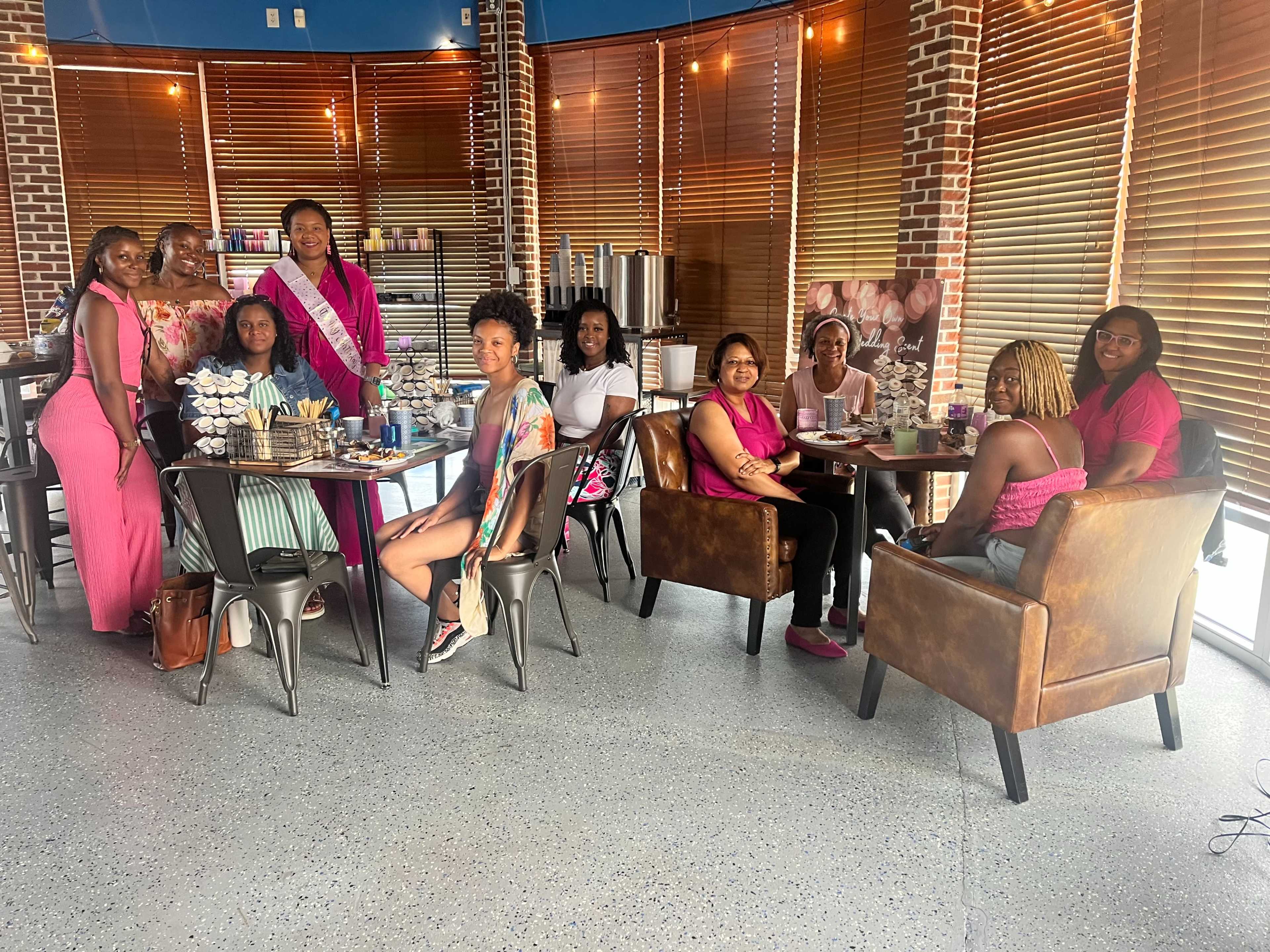 A group of nine women is gathered around tables in a brightly lit café with wooden blinds and modern furnishings.
